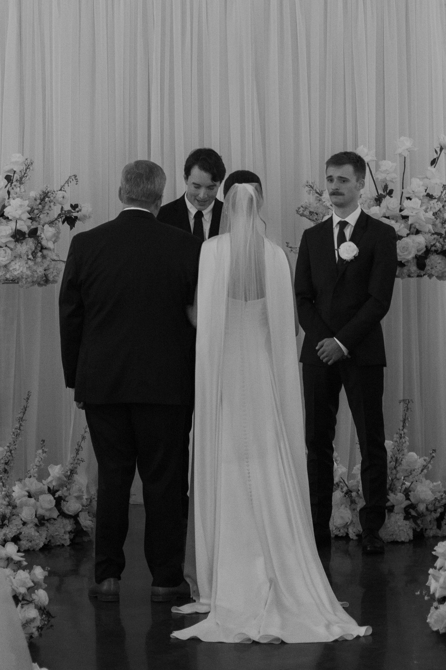 Wedding ceremony in progress with floral arrangements and white draping as backdrop.