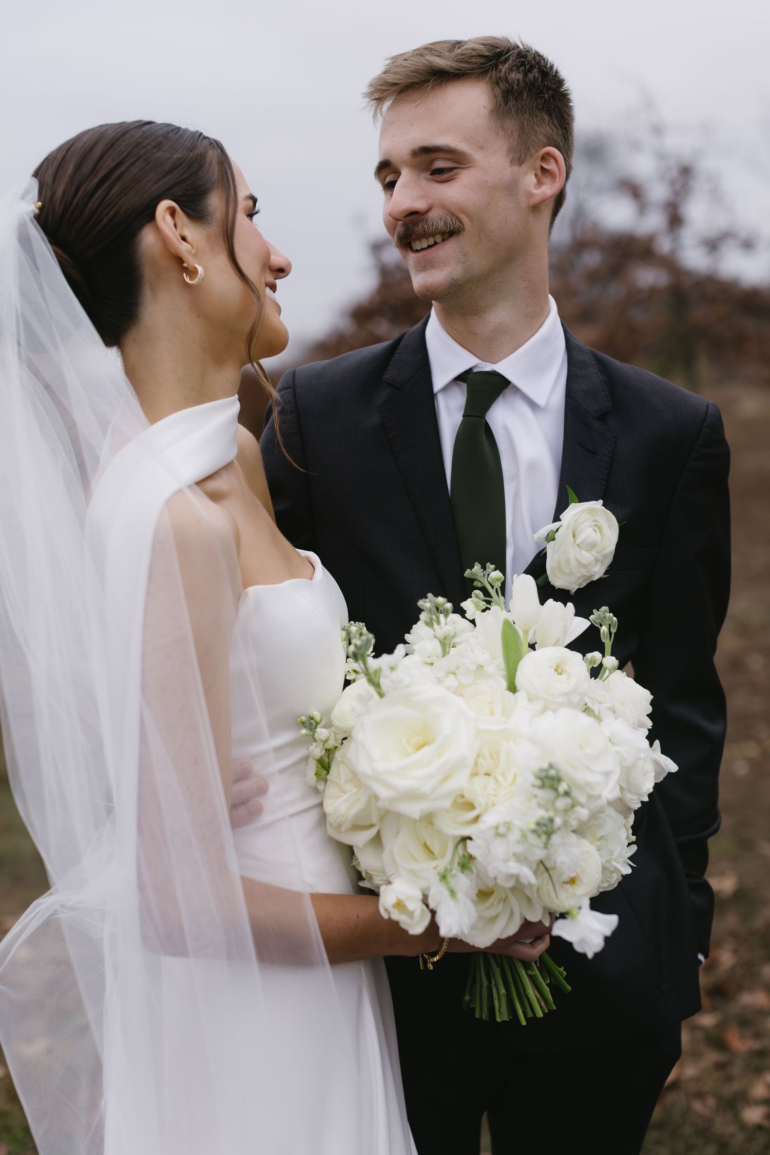 Bride in elegant white dress holds bouquet while sharing joyful moment with groom outdoors.