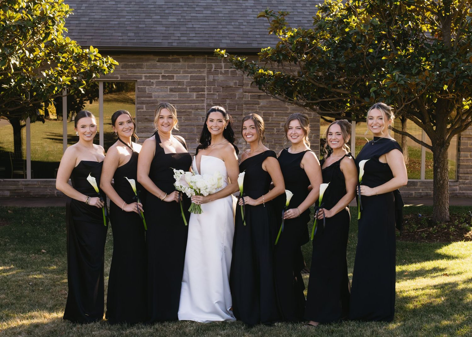 Wedding party poses outdoors with bridesmaids in black dresses against brick building.