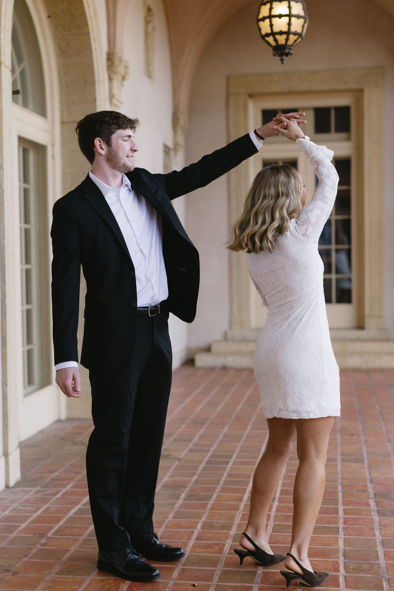 A couple dancing together in formal attire on a tiled porch with arched architecture and hanging lanterns.