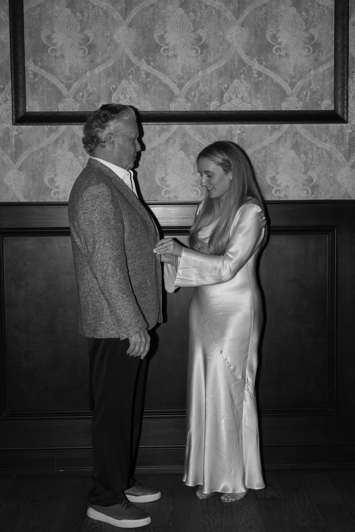 Black and white photo of a couple in formal wear sharing a tender moment against decorative wallpaper.