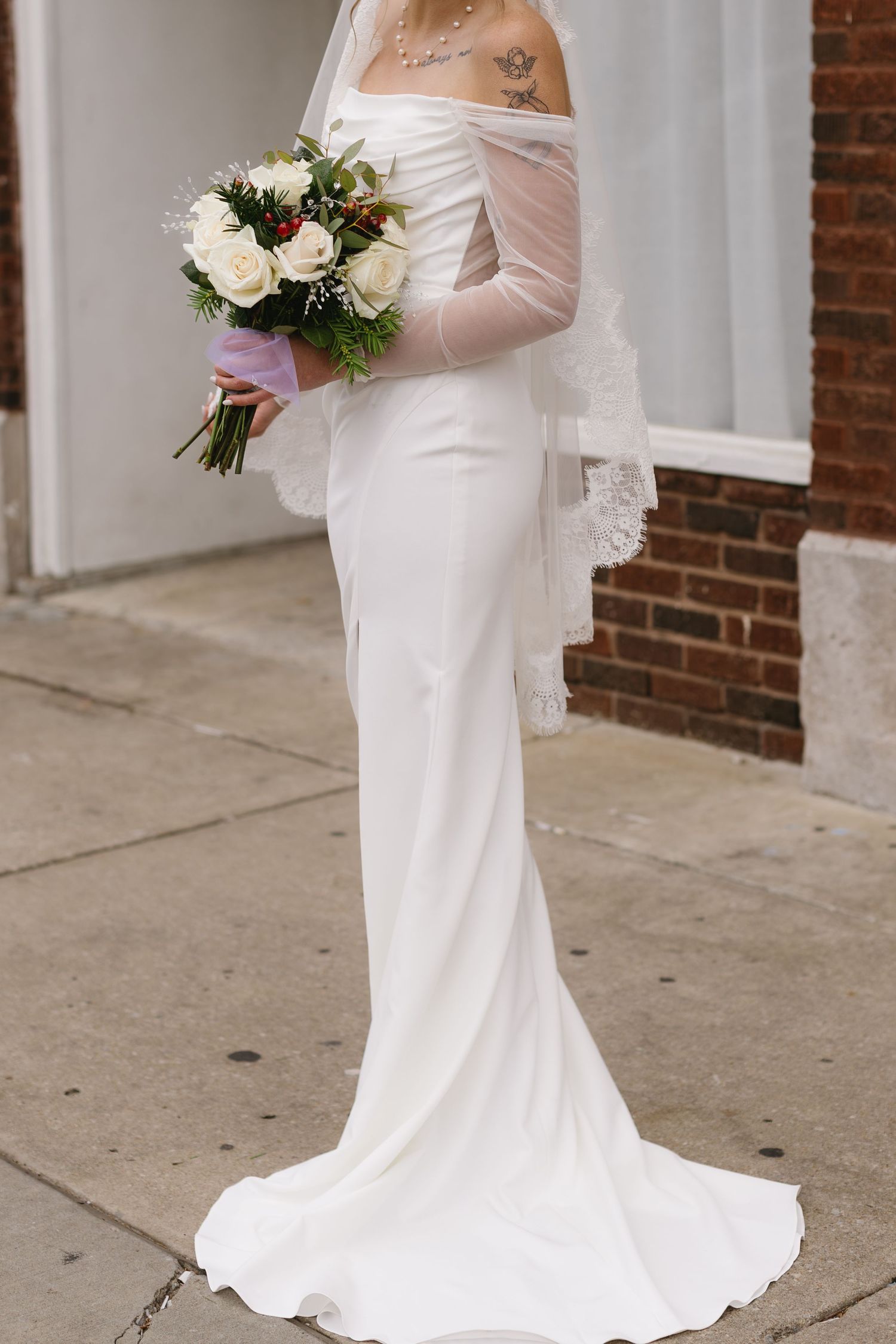 Close-up of bride in fitted white wedding gown holding floral bouquet against brick wall backdrop.