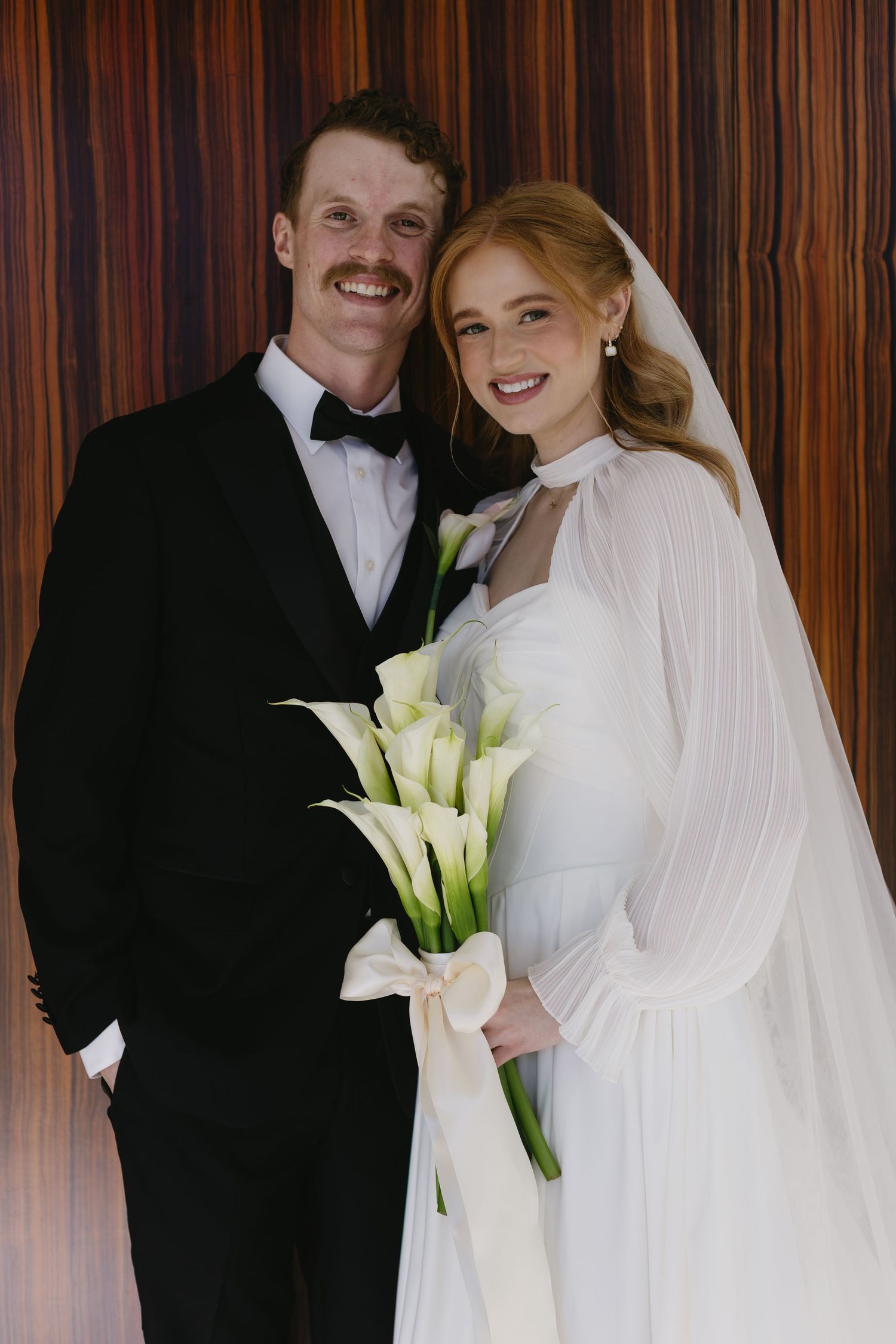 Wedding couple poses with white calla lily bouquet against wood paneled wall.
