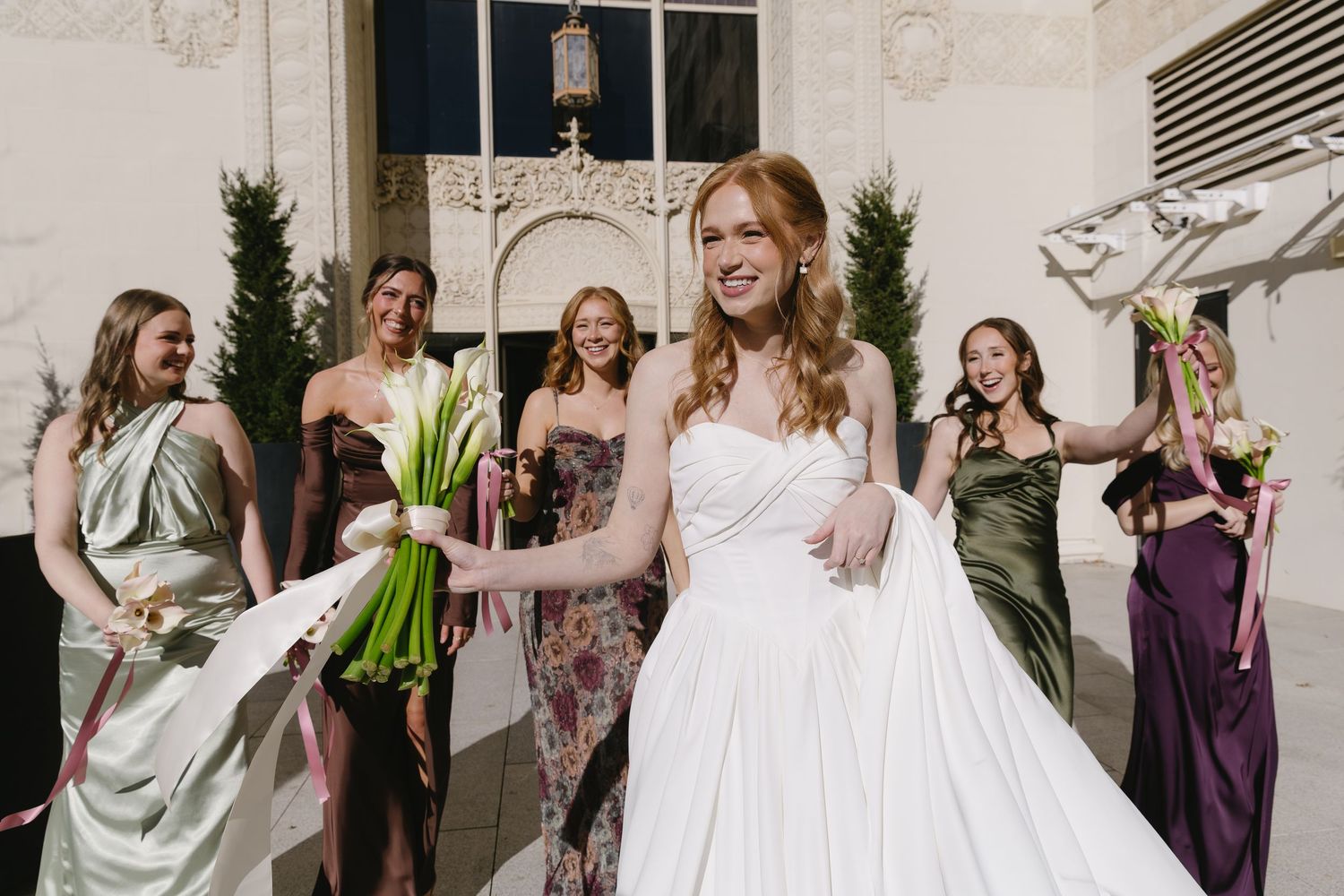 Wedding party in various colored dresses posing with calla lily bouquets outside a white building.
