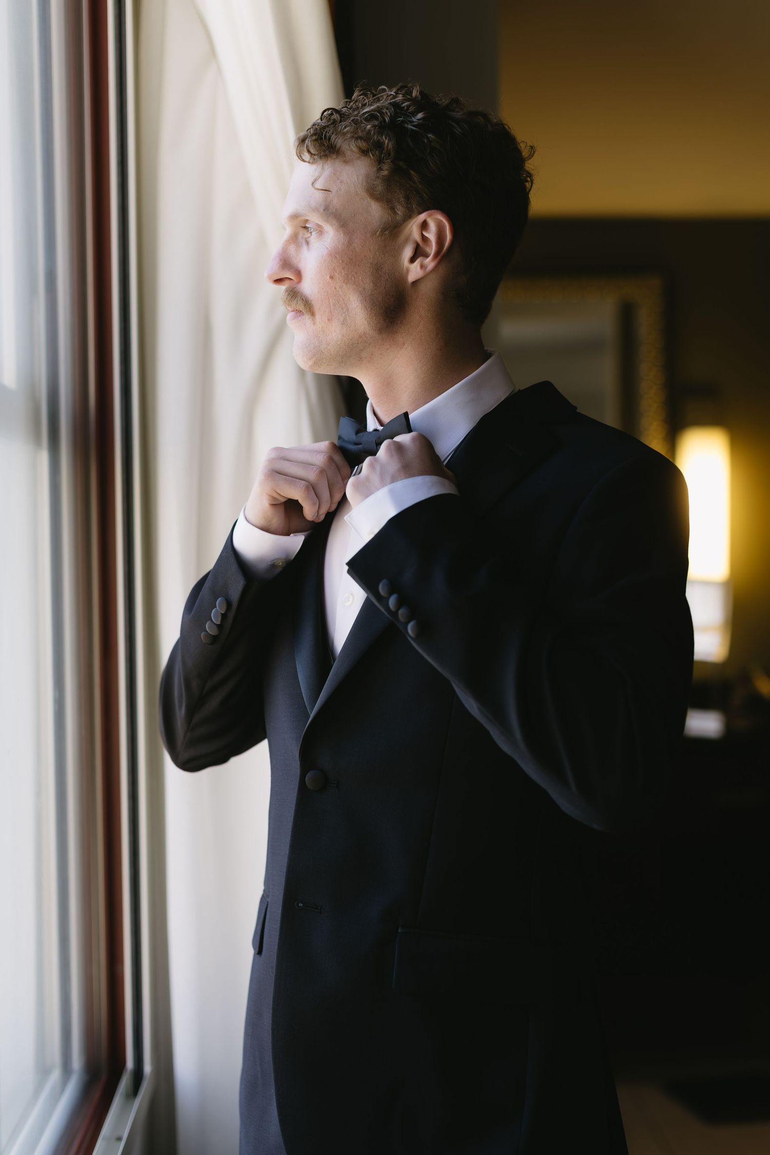 A man in a dark suit gazes thoughtfully out of a bright window in a contemplative pose.