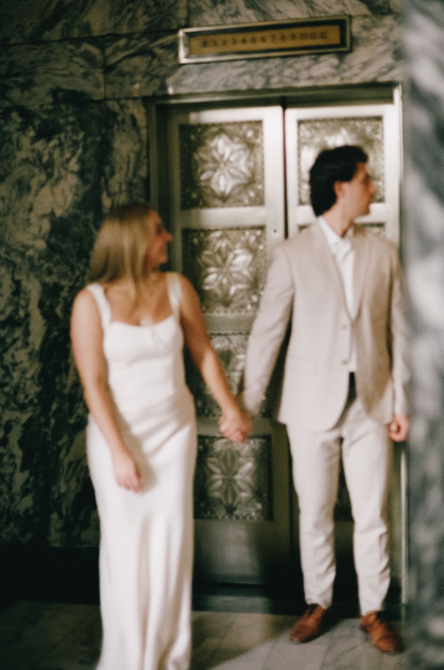A couple in matching white and beige outfits poses in an elegant vintage-style setting.