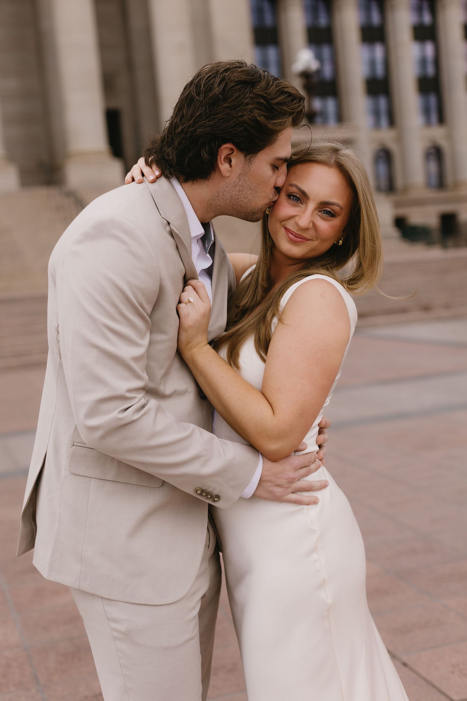 A romantic couple embraces and laughs together in front of an elegant stone facade.