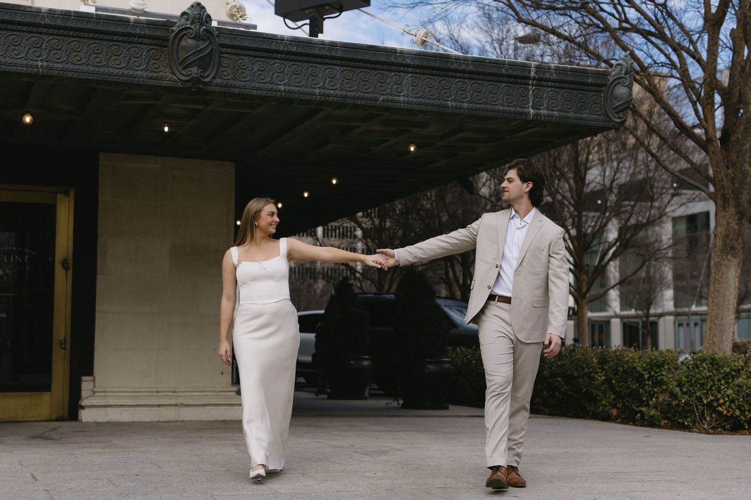 A couple in light colored formal wear poses playfully outdoors on a cloudy day.