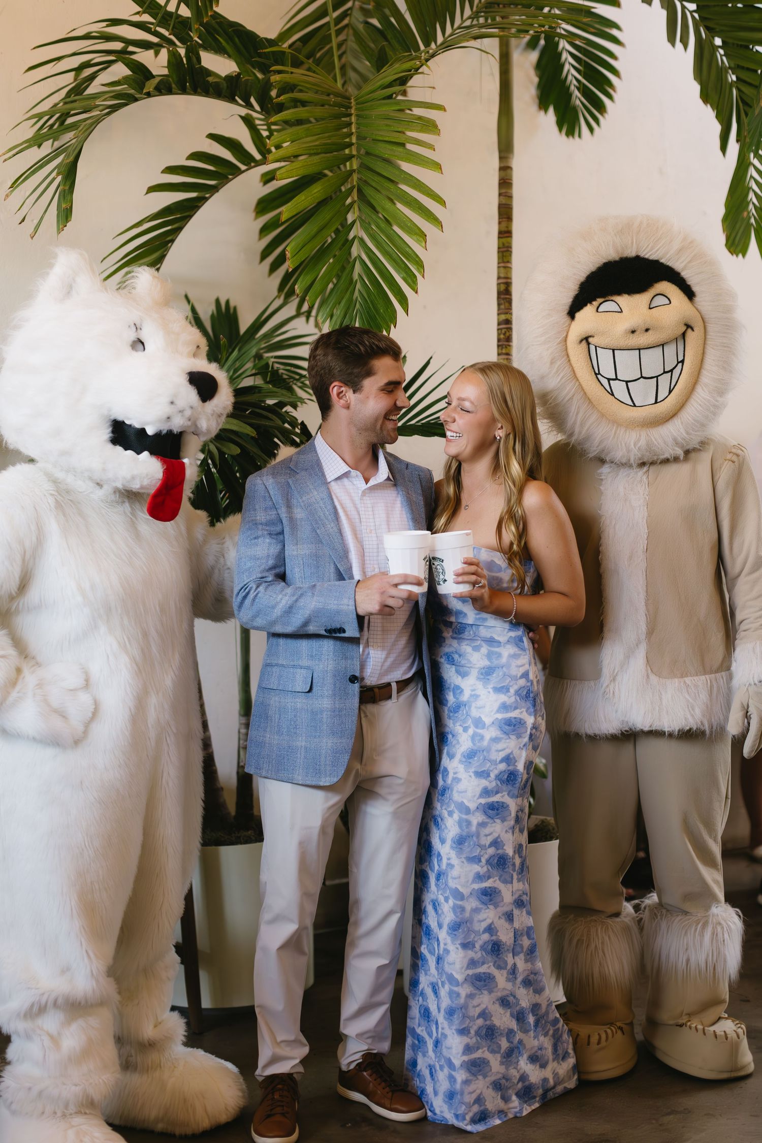 A couple poses with mascot characters under tropical plants at a special event.