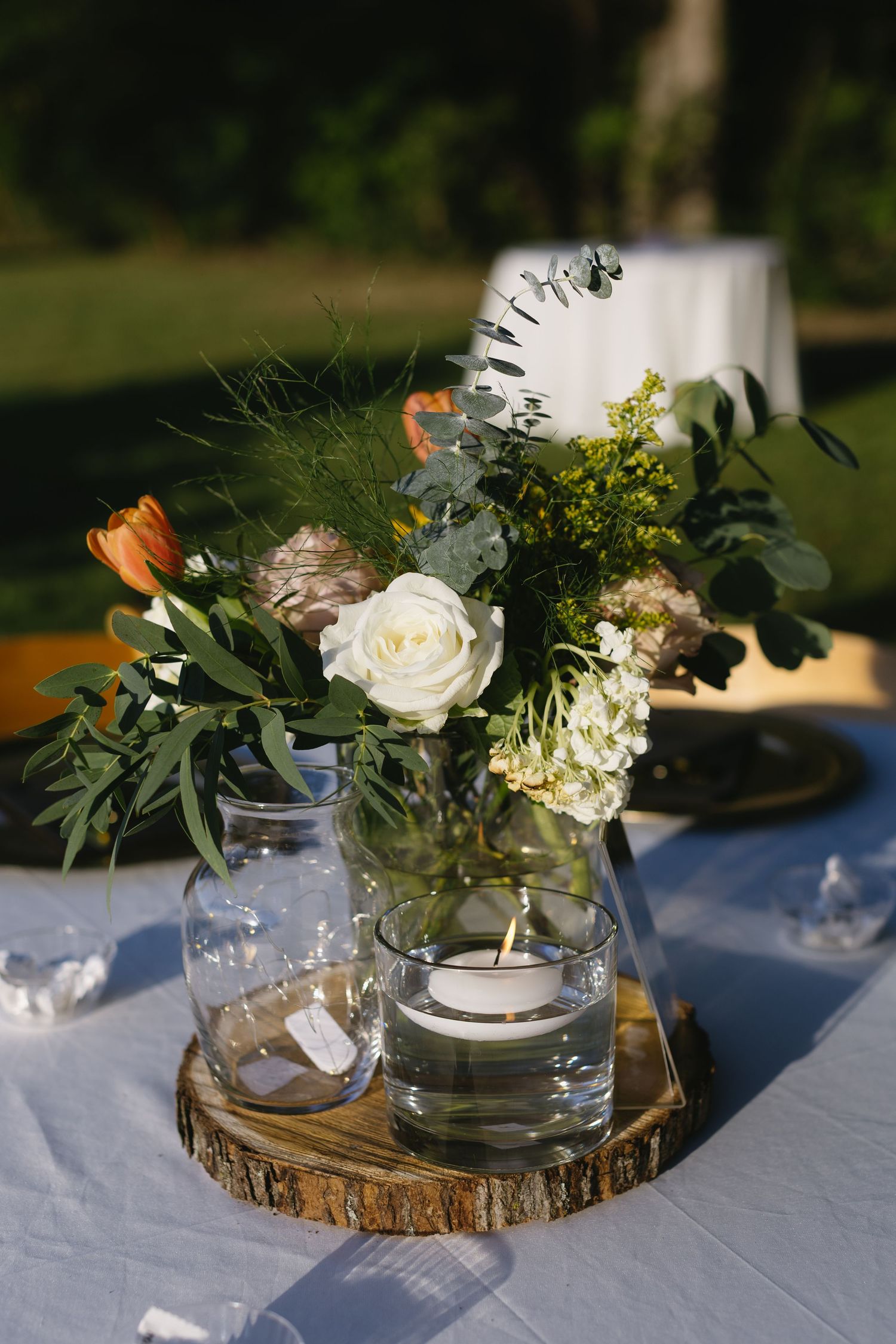 Wedding centerpieces with white roses and greenery arranged on wooden slices with glass vases.