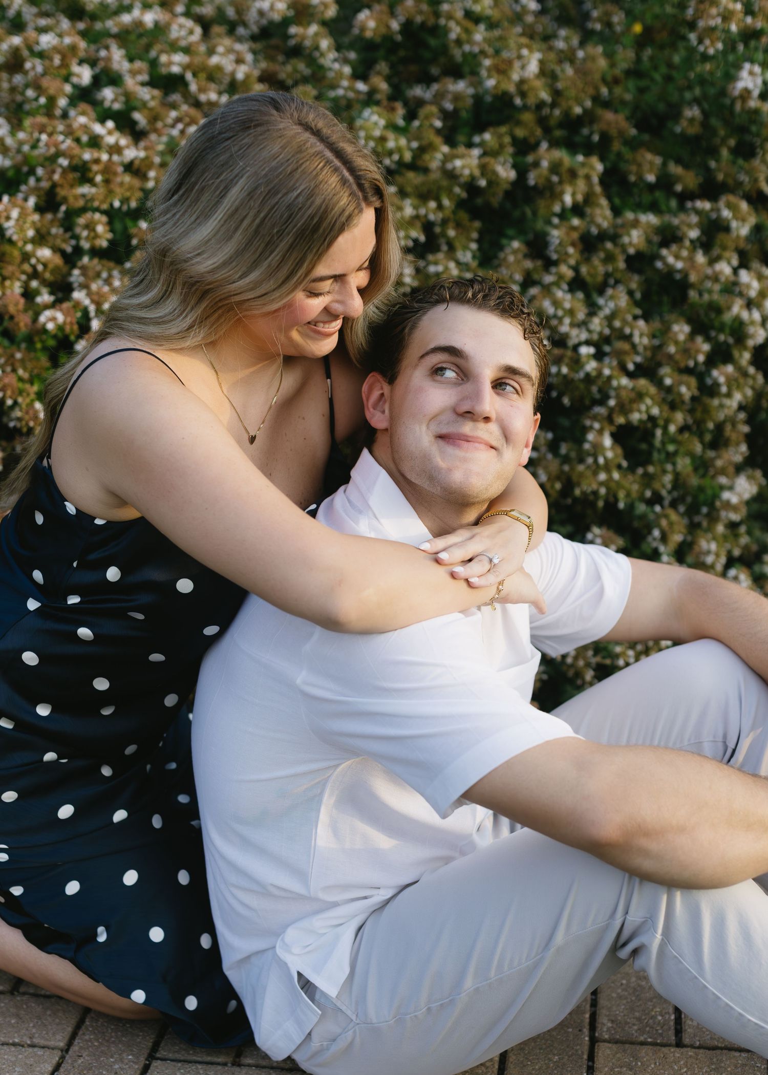 Couple relaxes together outdoors, sharing playful moment against flowering bush background.