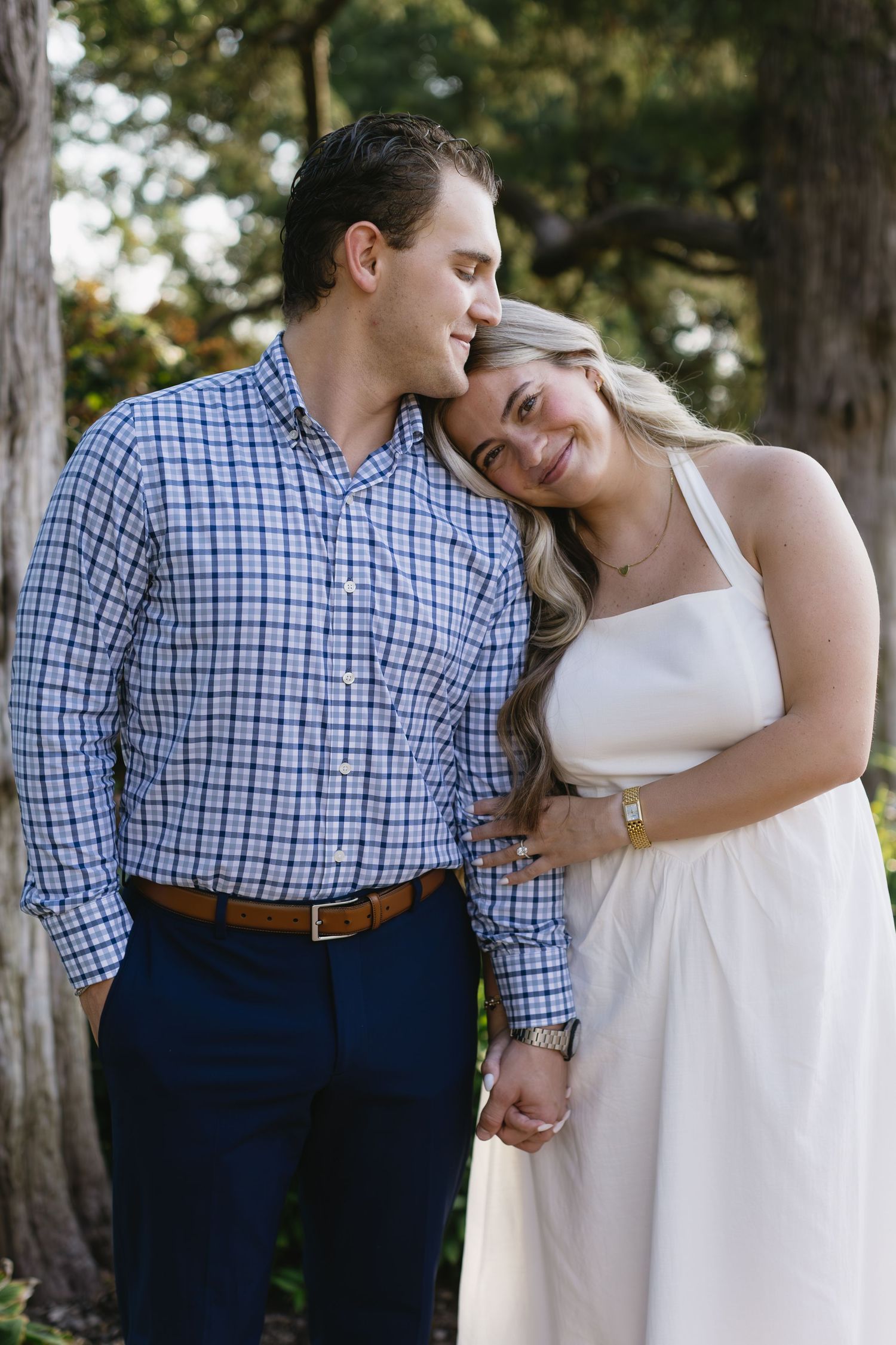 Couple shares an intimate moment outdoors as she leans on his shoulder wearing a white dress and he wears a plaid shirt.
