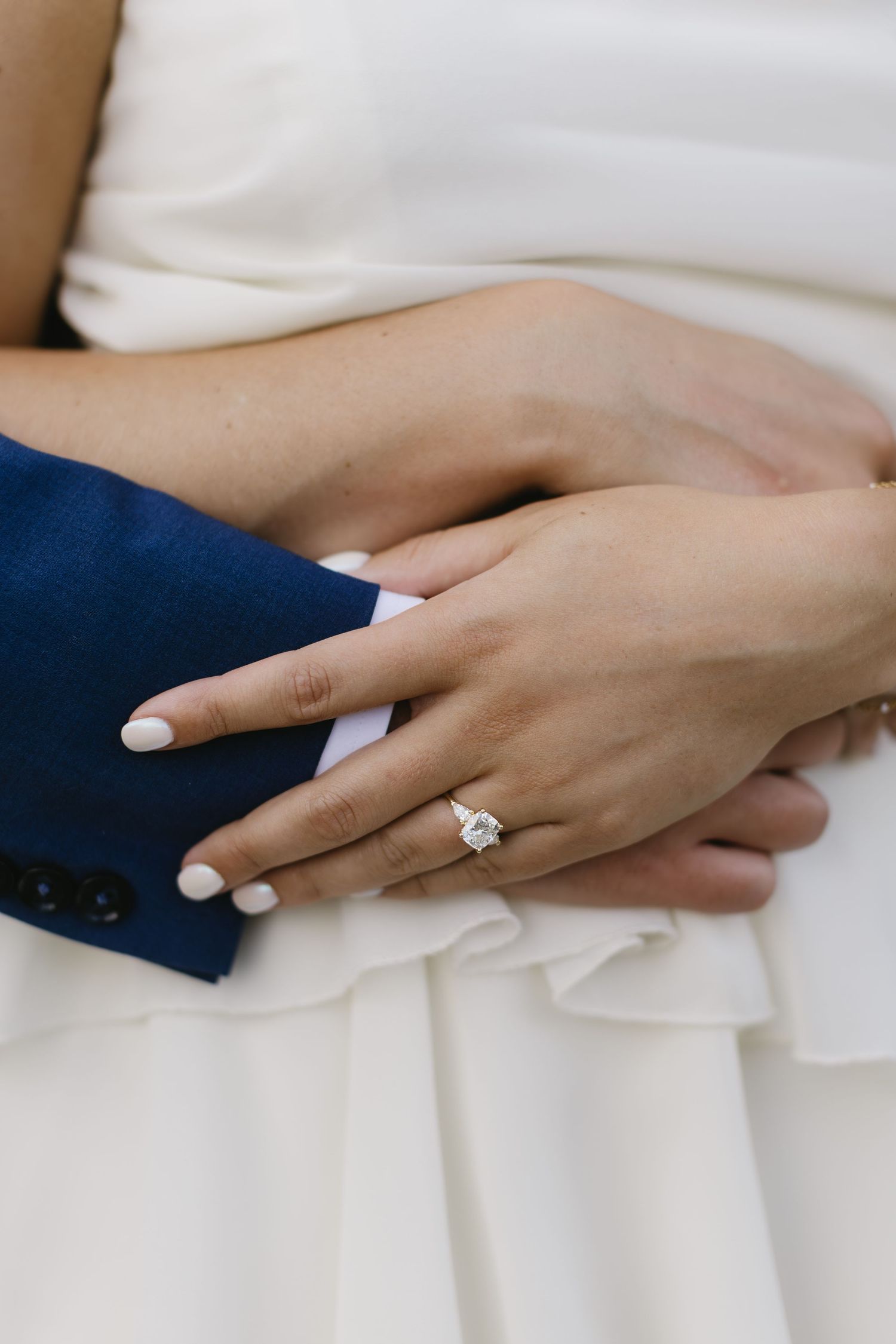 A hand wearing a sparkling diamond engagement ring rests delicately on white fabric.