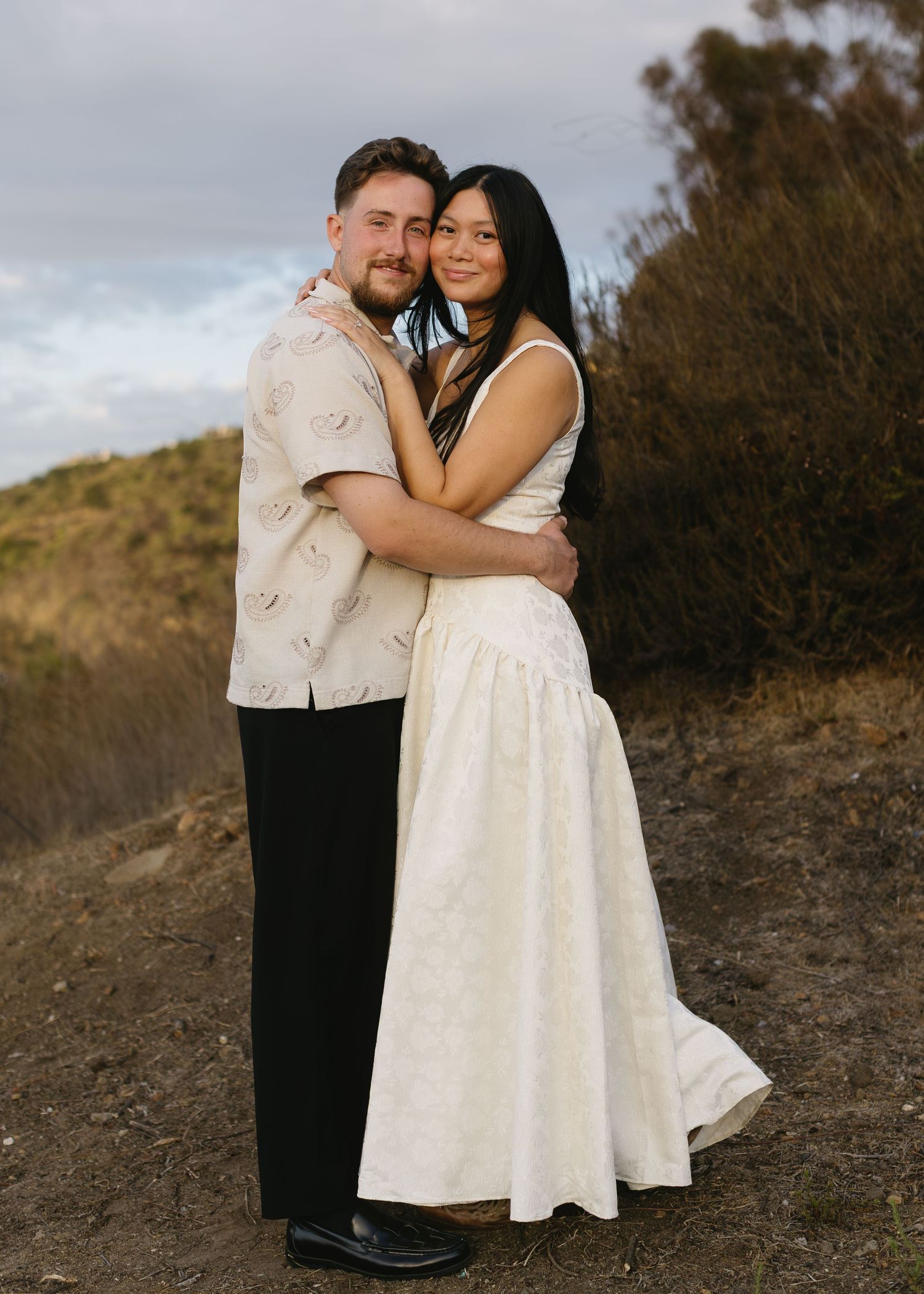 A couple poses together outdoors, with the person in white wearing a flowing dress against a natural backdrop.