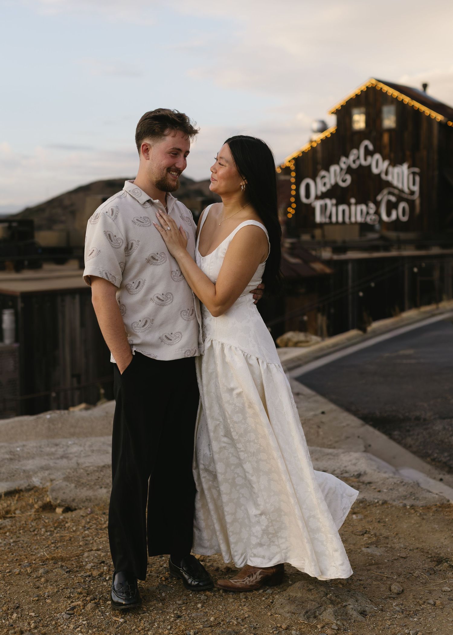 A romantic couple shares an intimate moment against a rustic wooden building backdrop at sunset.