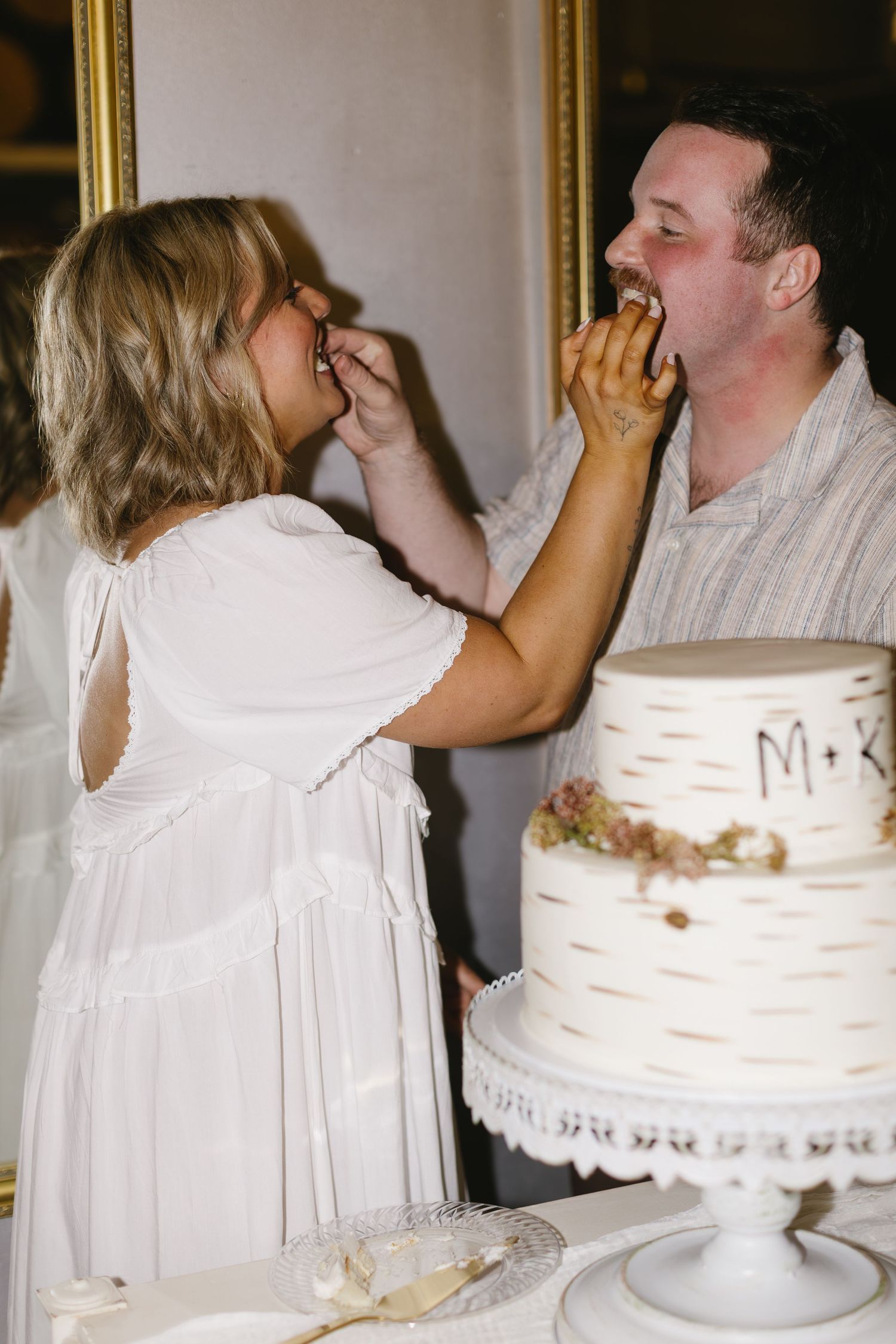 Newlyweds playfully feed each other wedding cake beside their white birch-themed tiered dessert.