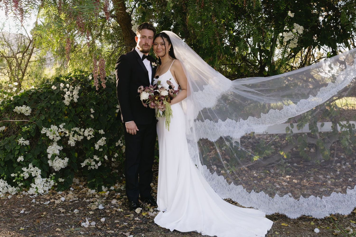 A bride in a long veil poses with her groom against a lush garden backdrop.