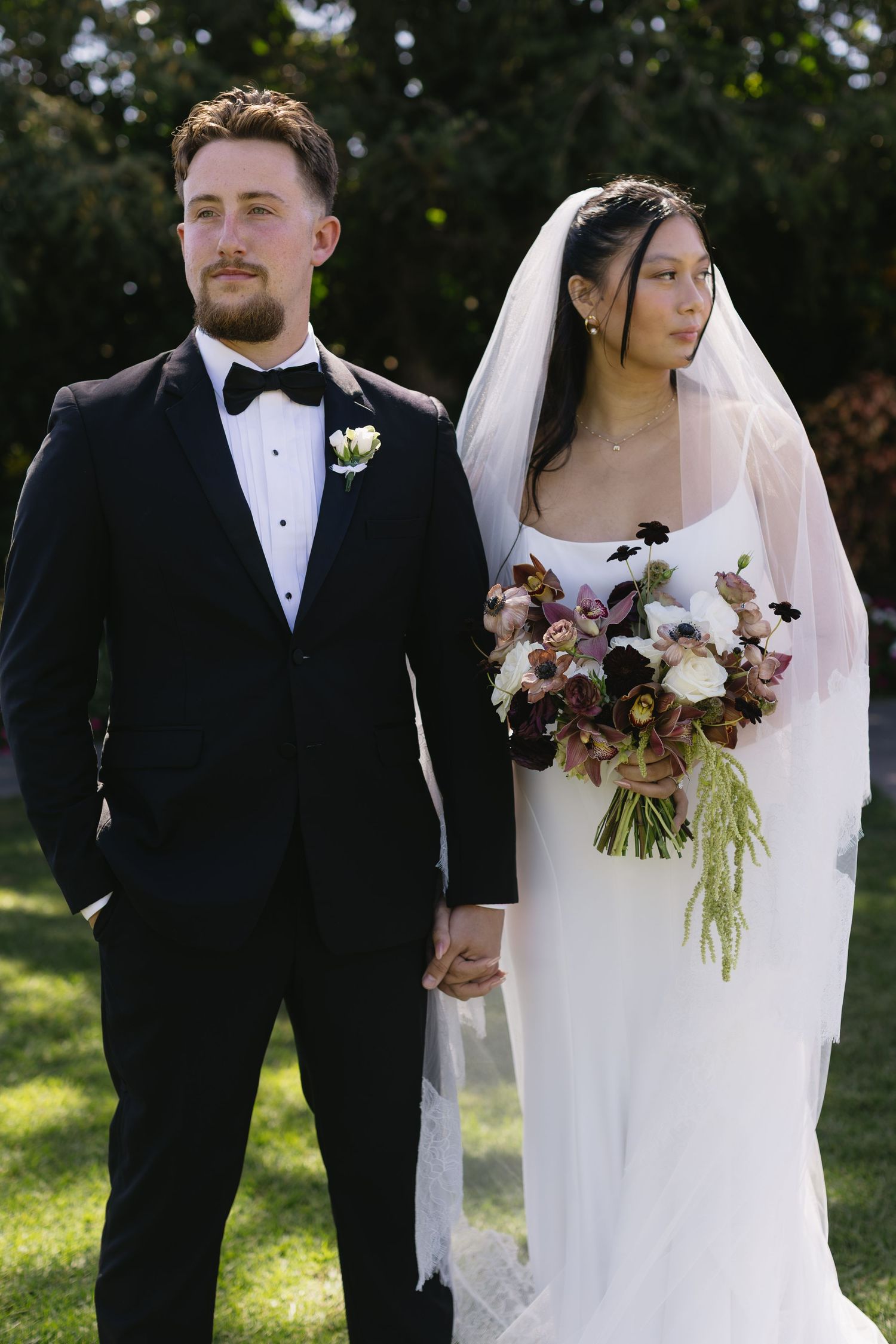 Newlywed couple poses outdoors with bride holding dramatic burgundy and white bouquet.