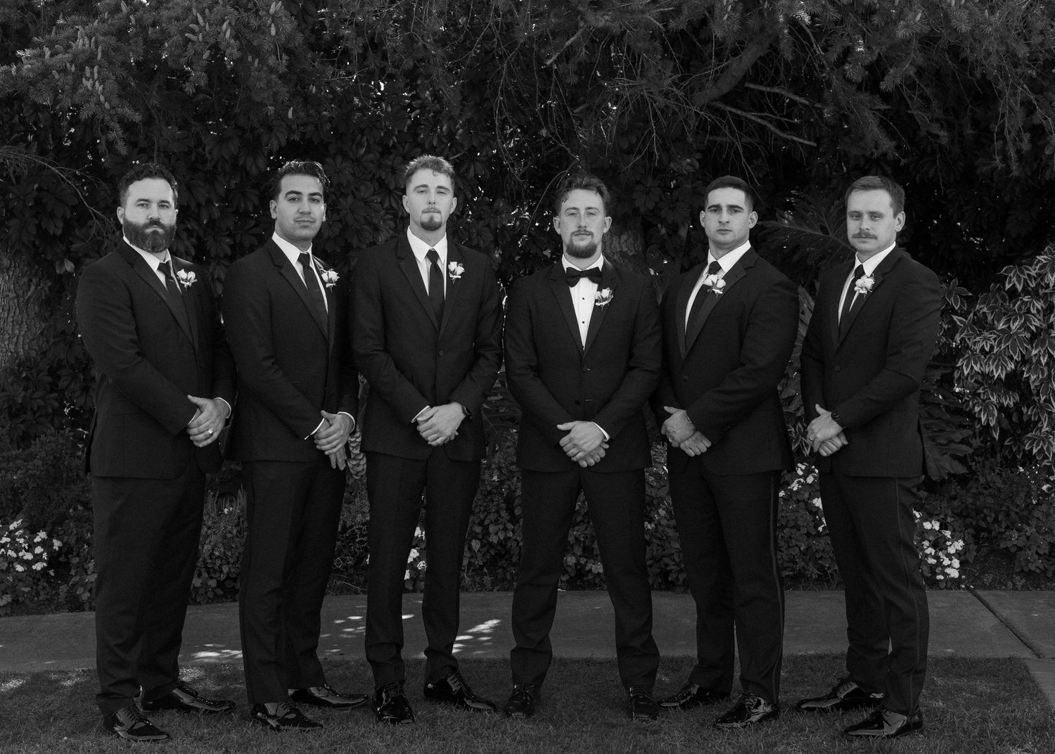 Groomsmen in black tuxedos lined up for formal wedding photos outdoors.