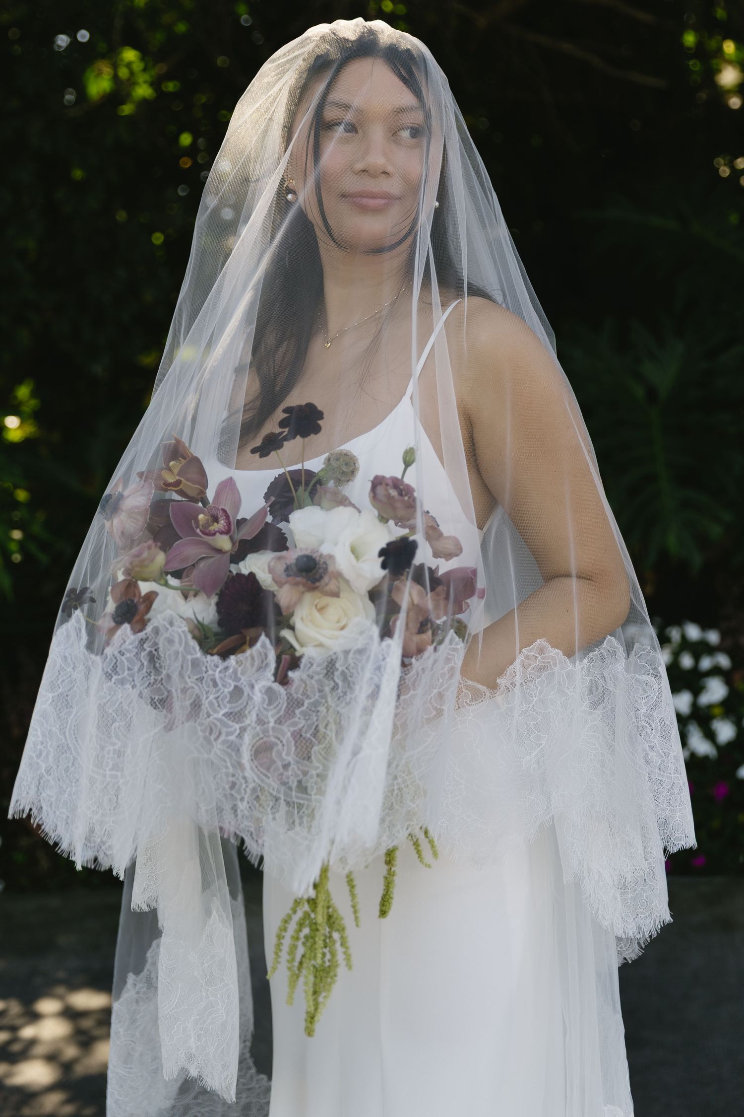 A bride wearing a delicate lace veil with floral details stands against a natural outdoor backdrop.