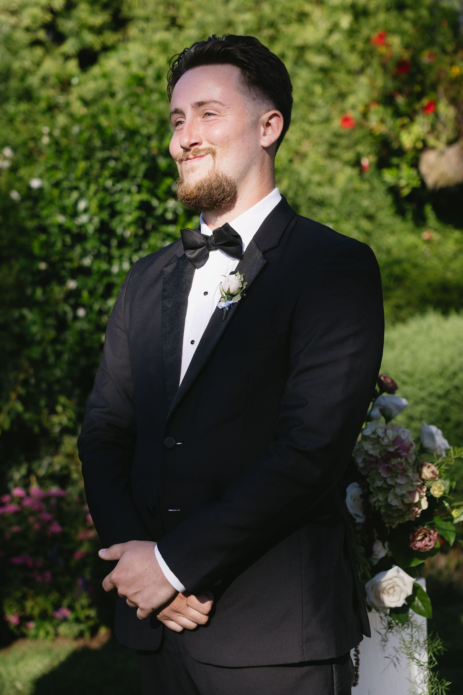 Groom in black tuxedo smiles during outdoor wedding ceremony.
