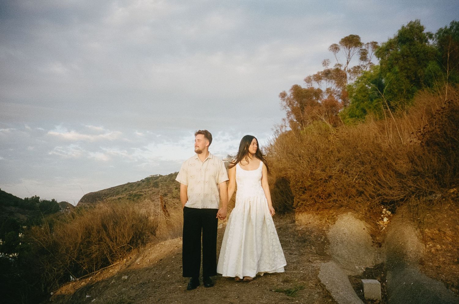 A couple stands together on a scenic hillside trail during golden hour sunset.