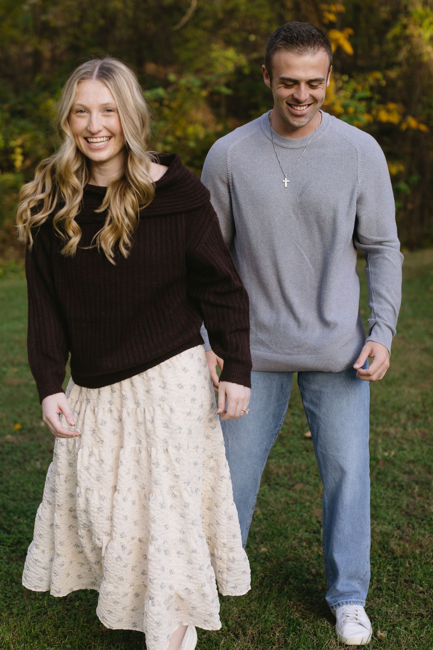 A couple in casual attire poses together outdoors during a fall photo session on green grass.
