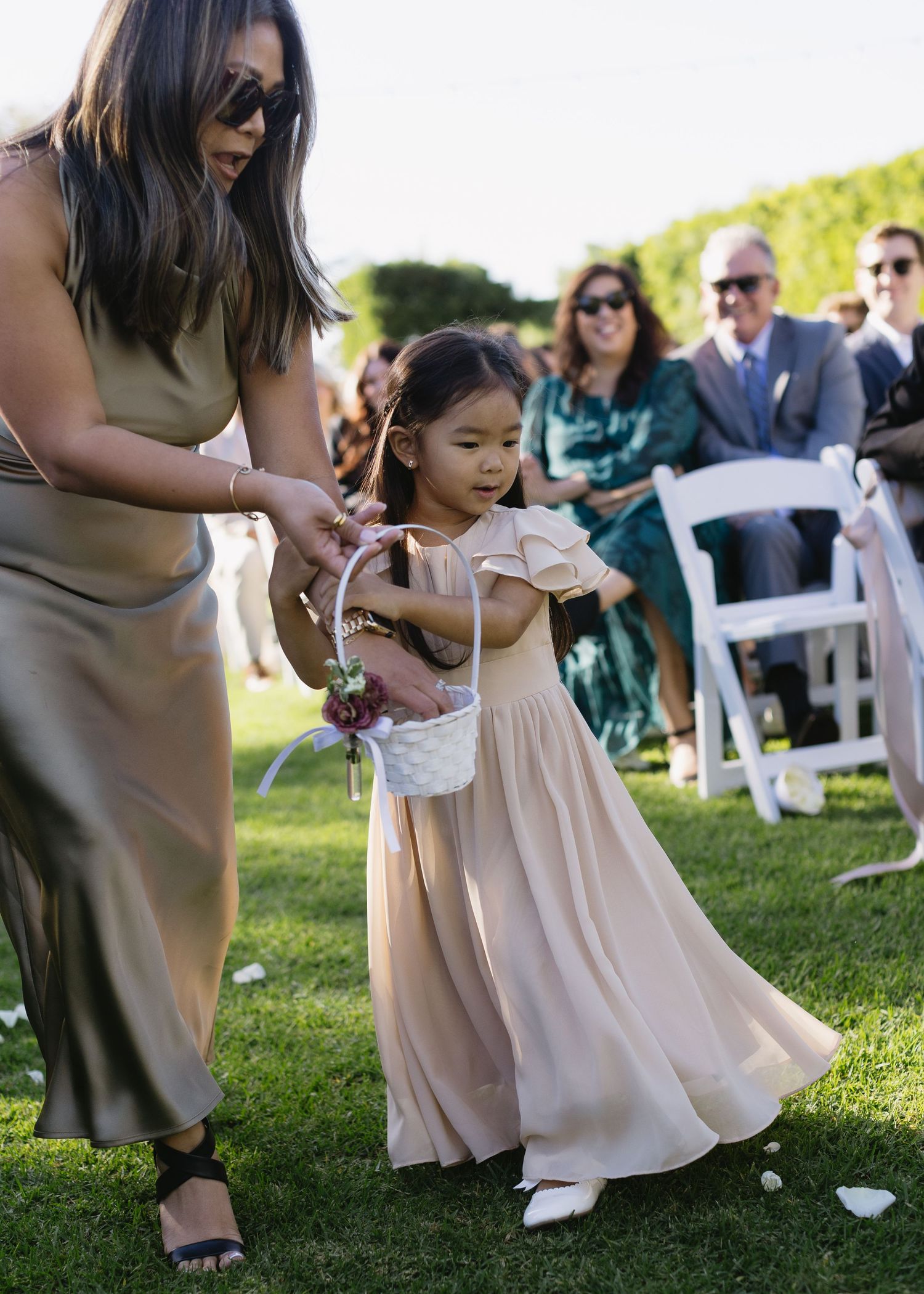 Child in a light pink dress twirls on grassy lawn during outdoor wedding ceremony.