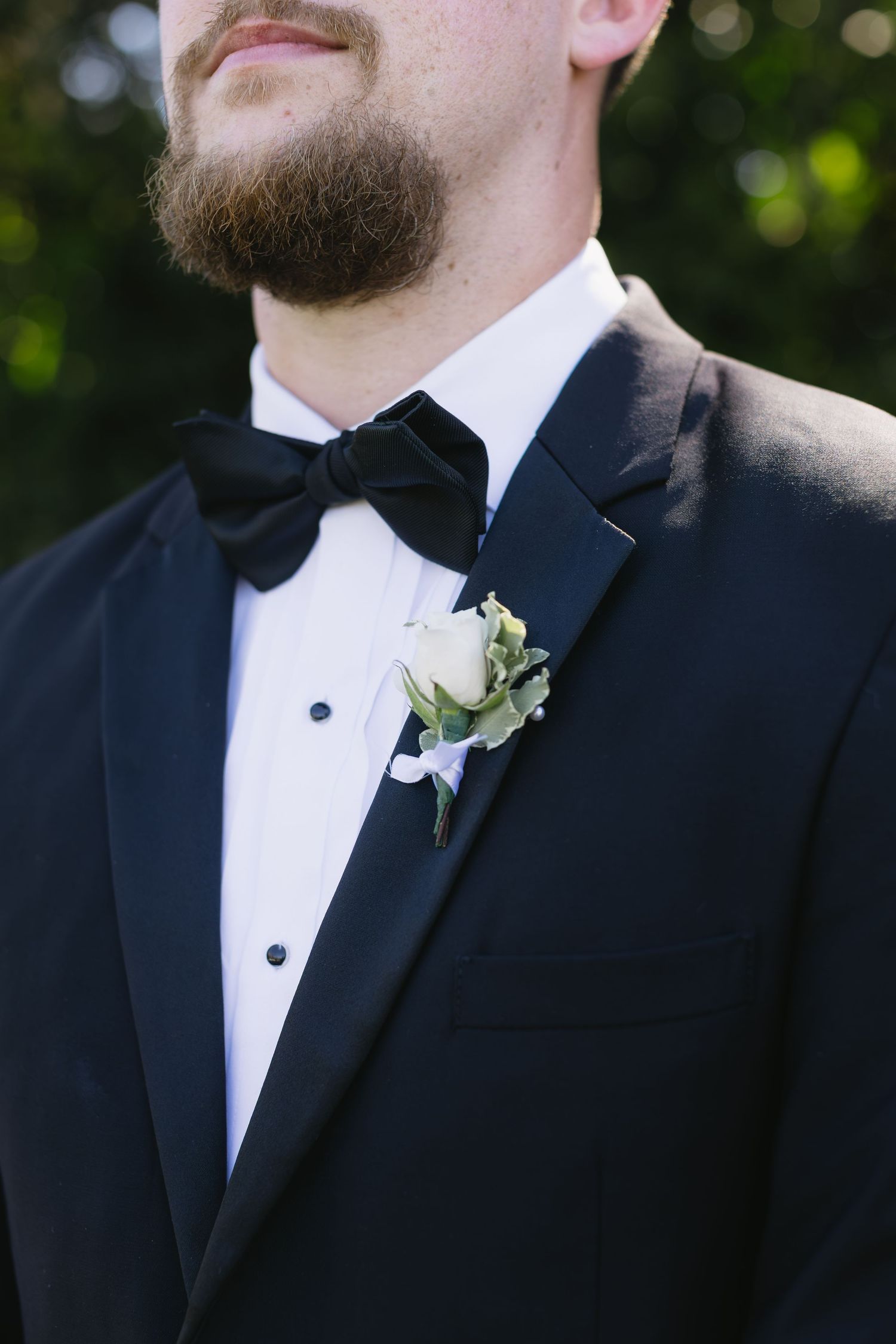 Close-up details of groomsmen in classic black tuxedos with white boutonnieres against a blurred outdoor background.