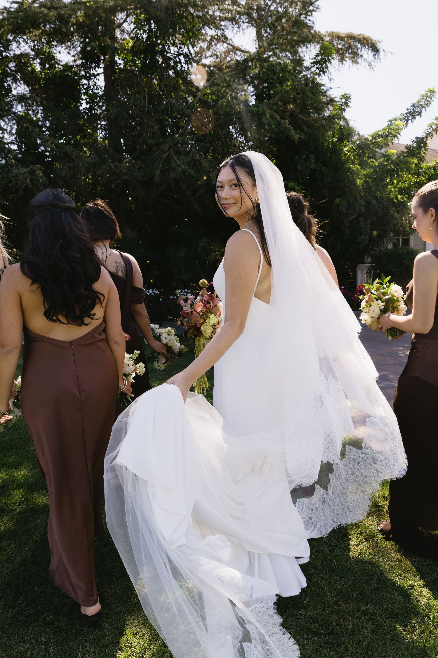 Wedding party in brown dresses and white gown walking together on a sunny outdoor lawn.