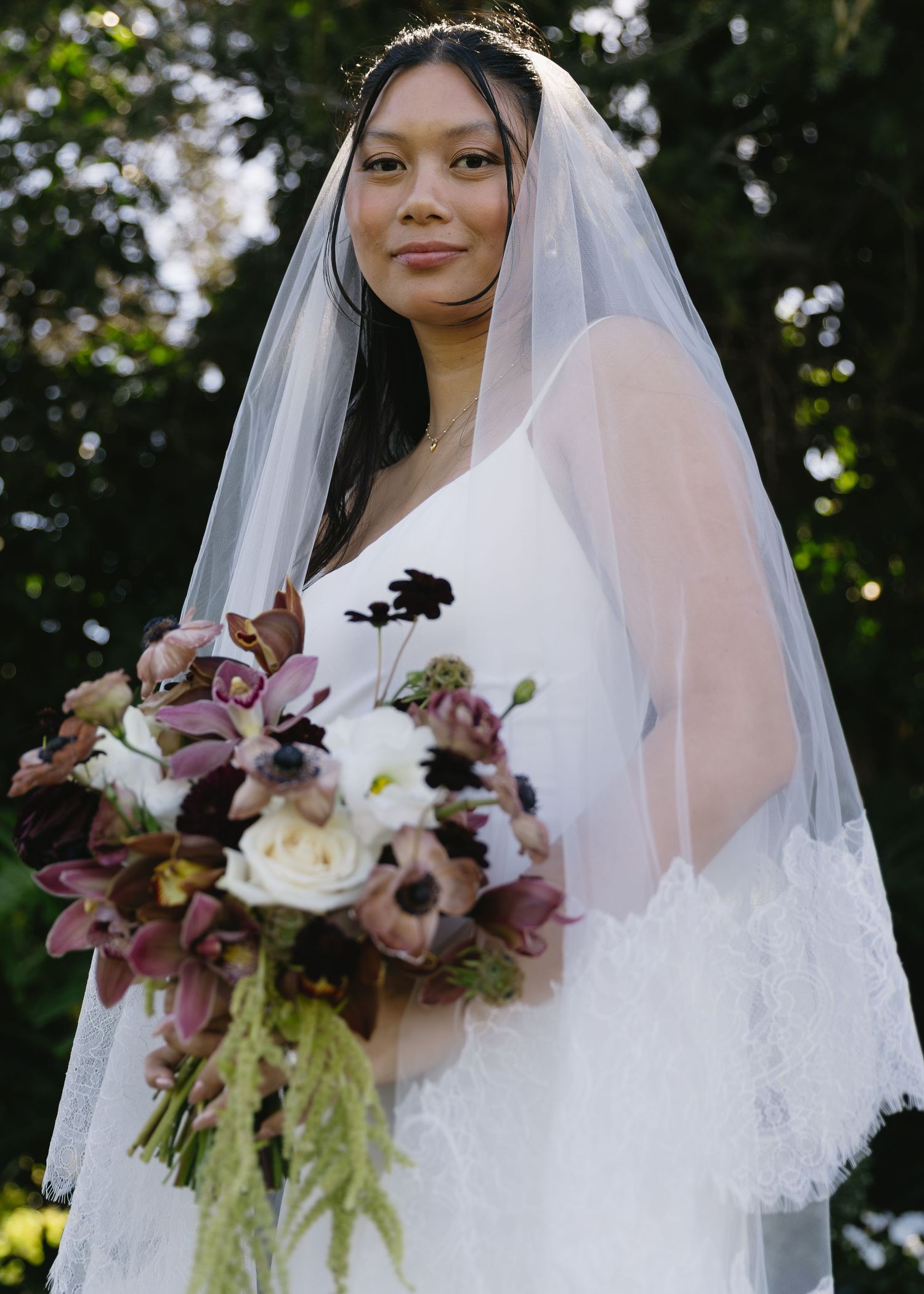 A bride in a lace veil holds an elegant bouquet with burgundy flowers and cascading greenery.