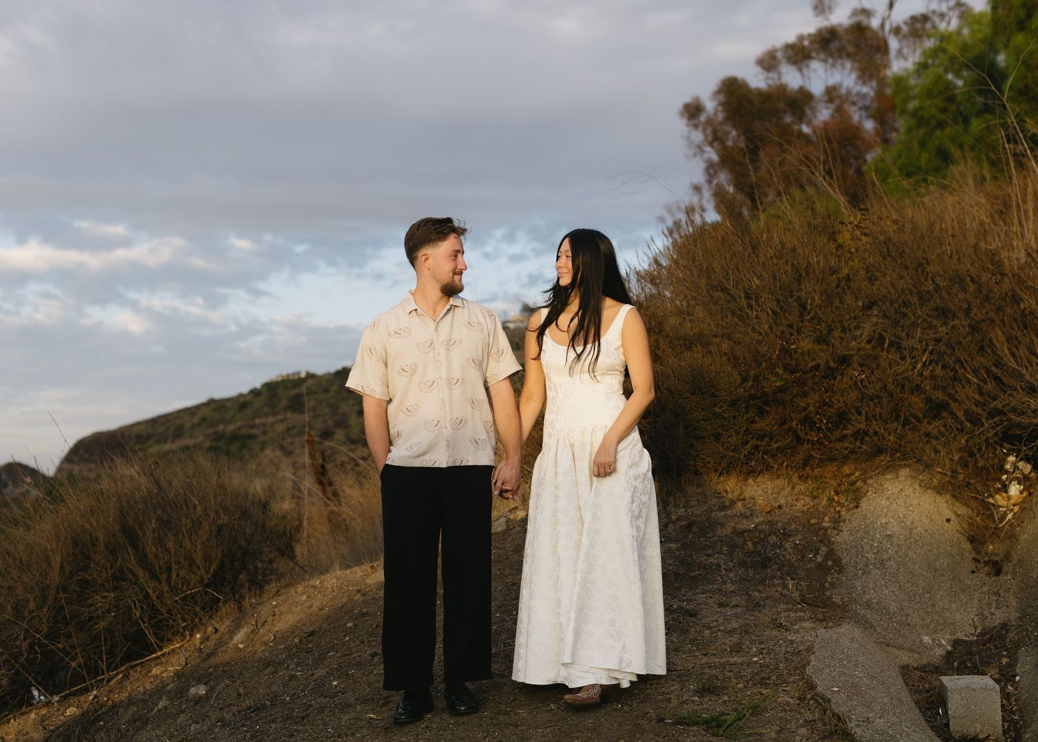 A romantic couple poses together on a hillside path during golden hour at sunset.