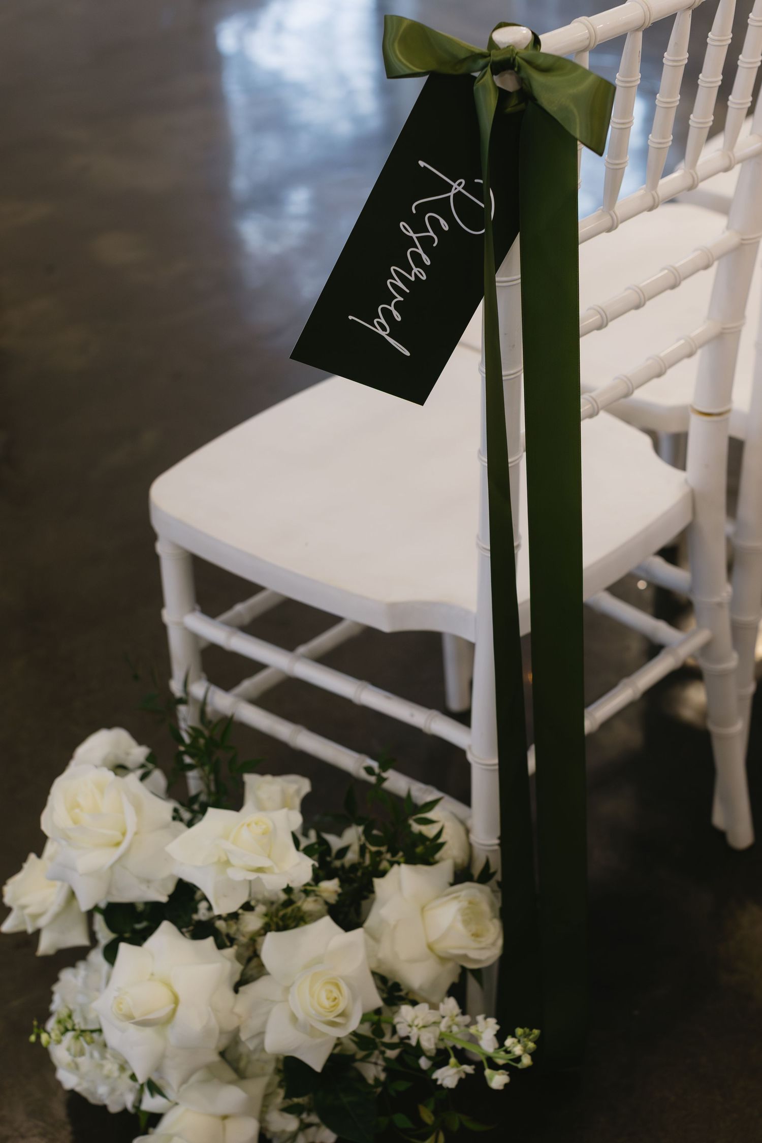 White ceremony chairs decorated with dark green ribbons and white roses for an elegant wedding.