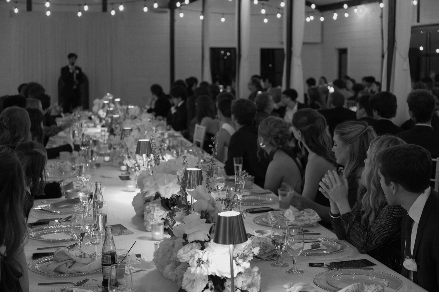 Black and white photo of elegant wedding reception with long tables and string lights.