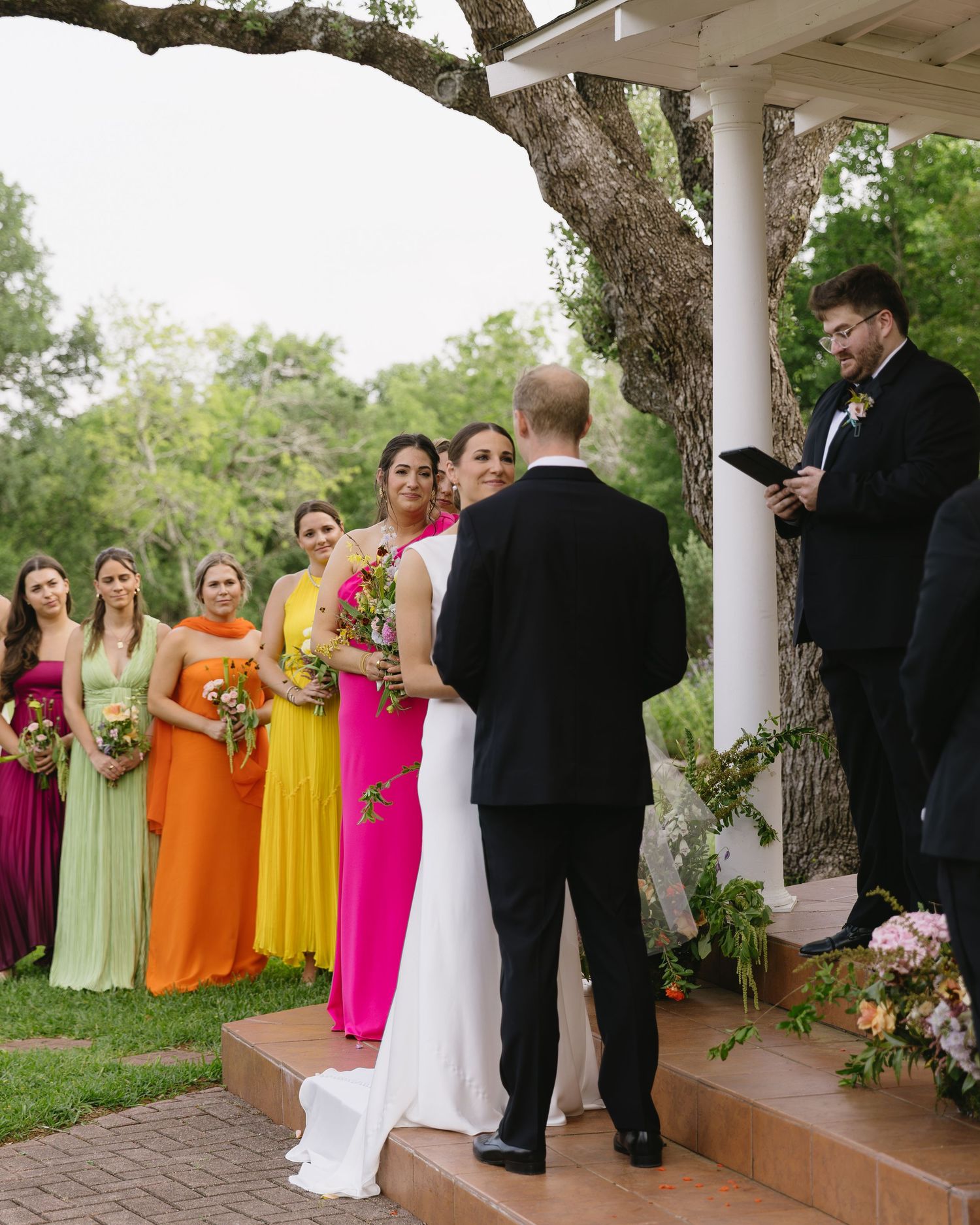 Outdoor wedding ceremony with bridesmaids in colorful dresses standing under tree.
