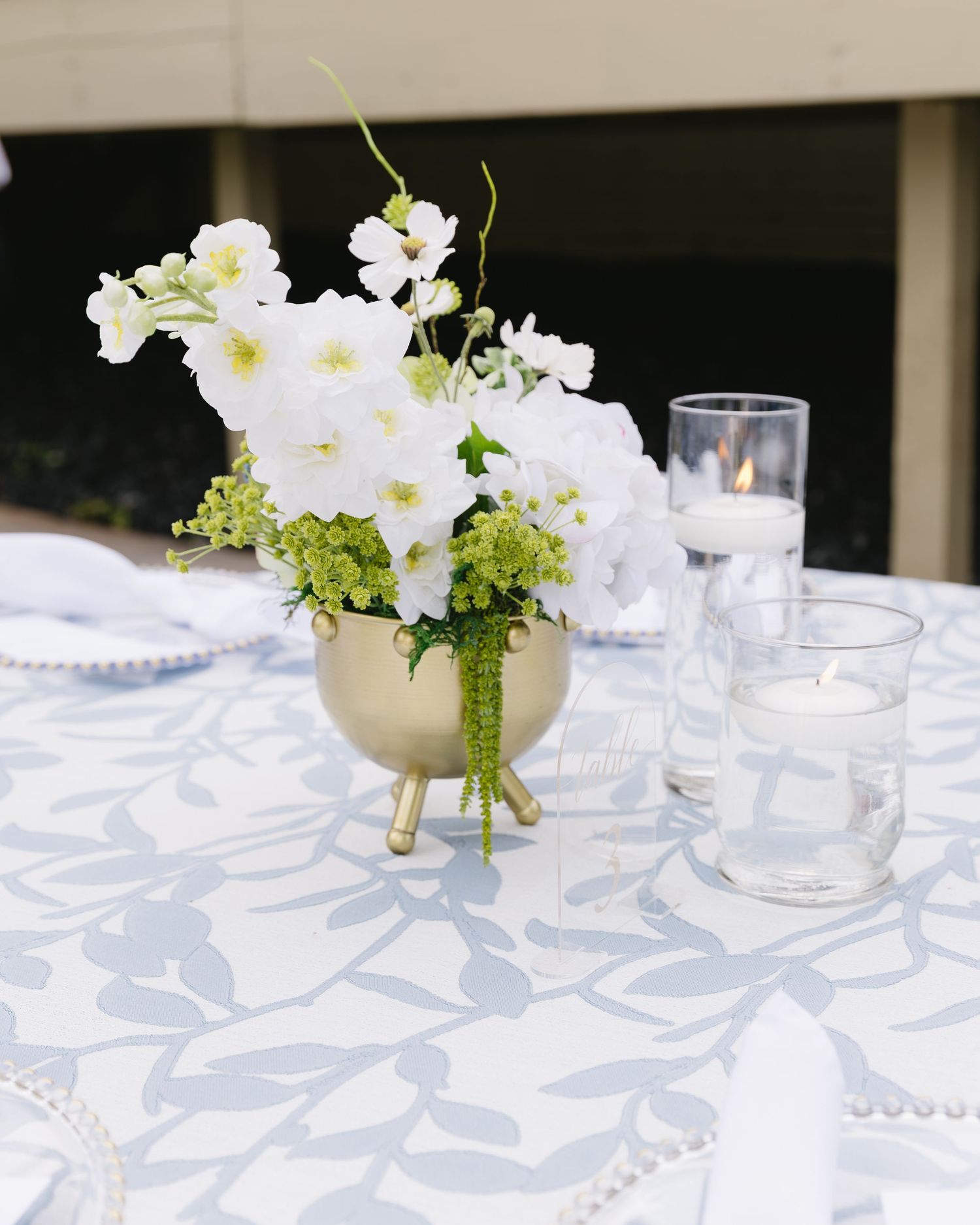 White floral centerpiece with orchids in a gold vase on blue patterned tablecloth.