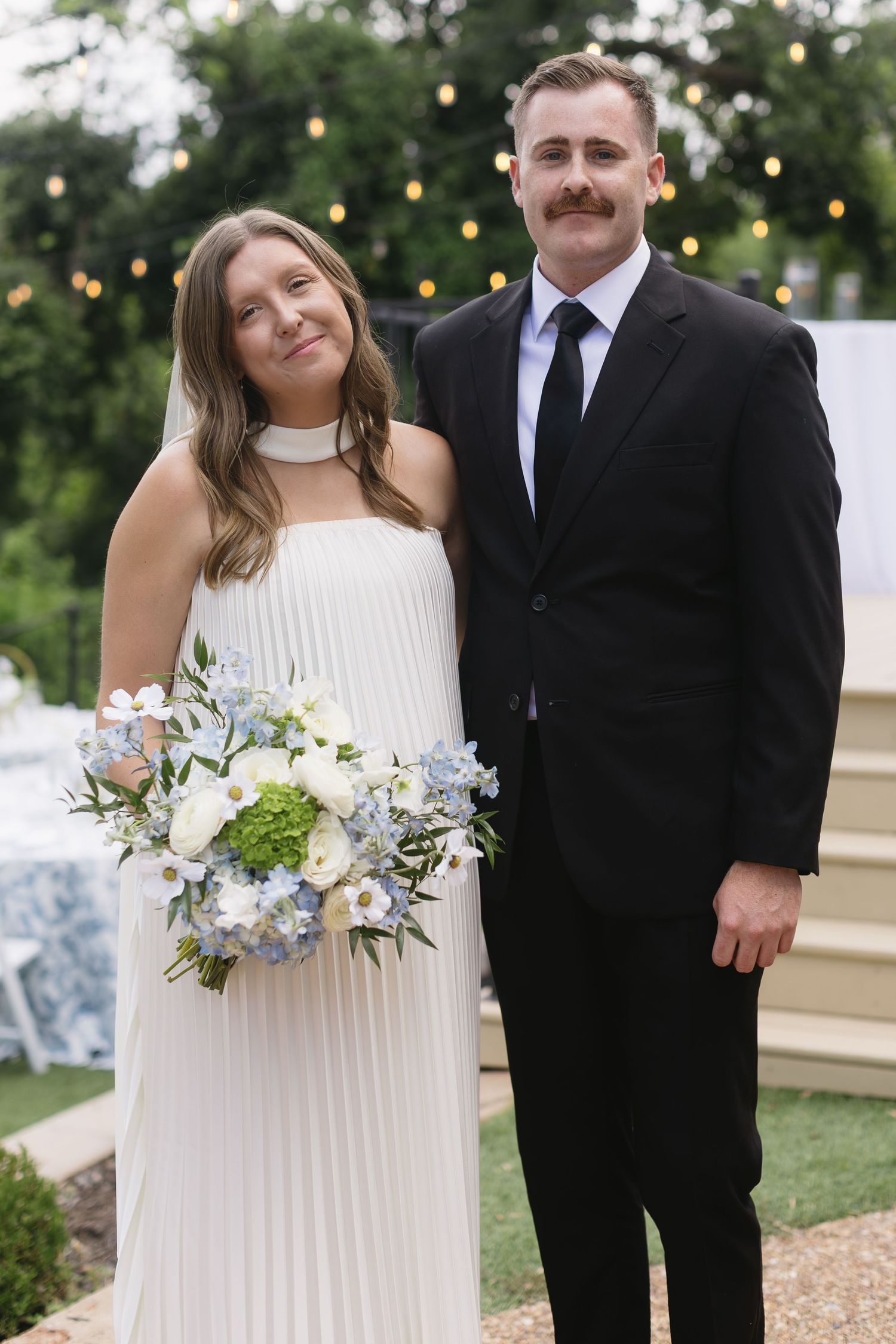 Couple in formal wedding attire standing outdoors with string lights and greenery in background.