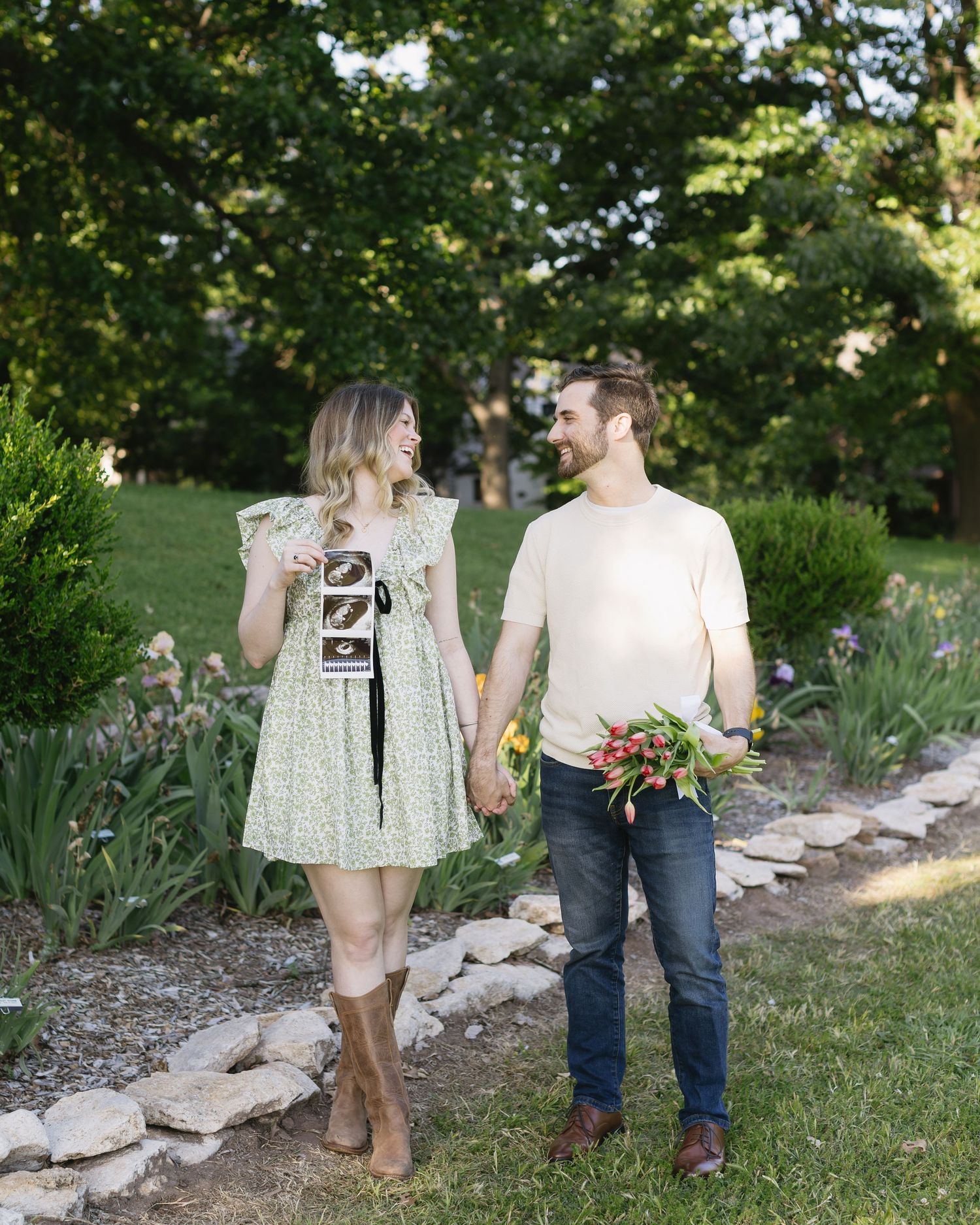 A couple holding ultrasound photos walks together in a garden path lined with rocks and greenery.