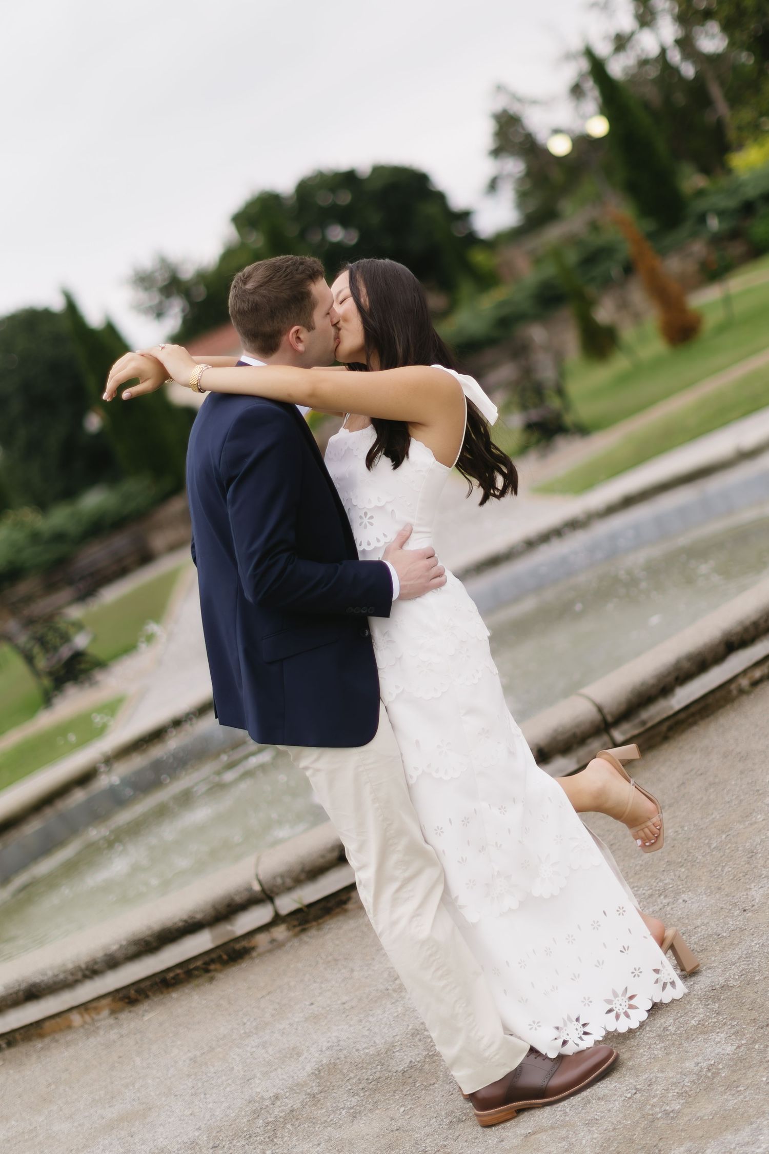 Couple in formal attire embrace in romantic outdoor setting with white dress and navy blazer.