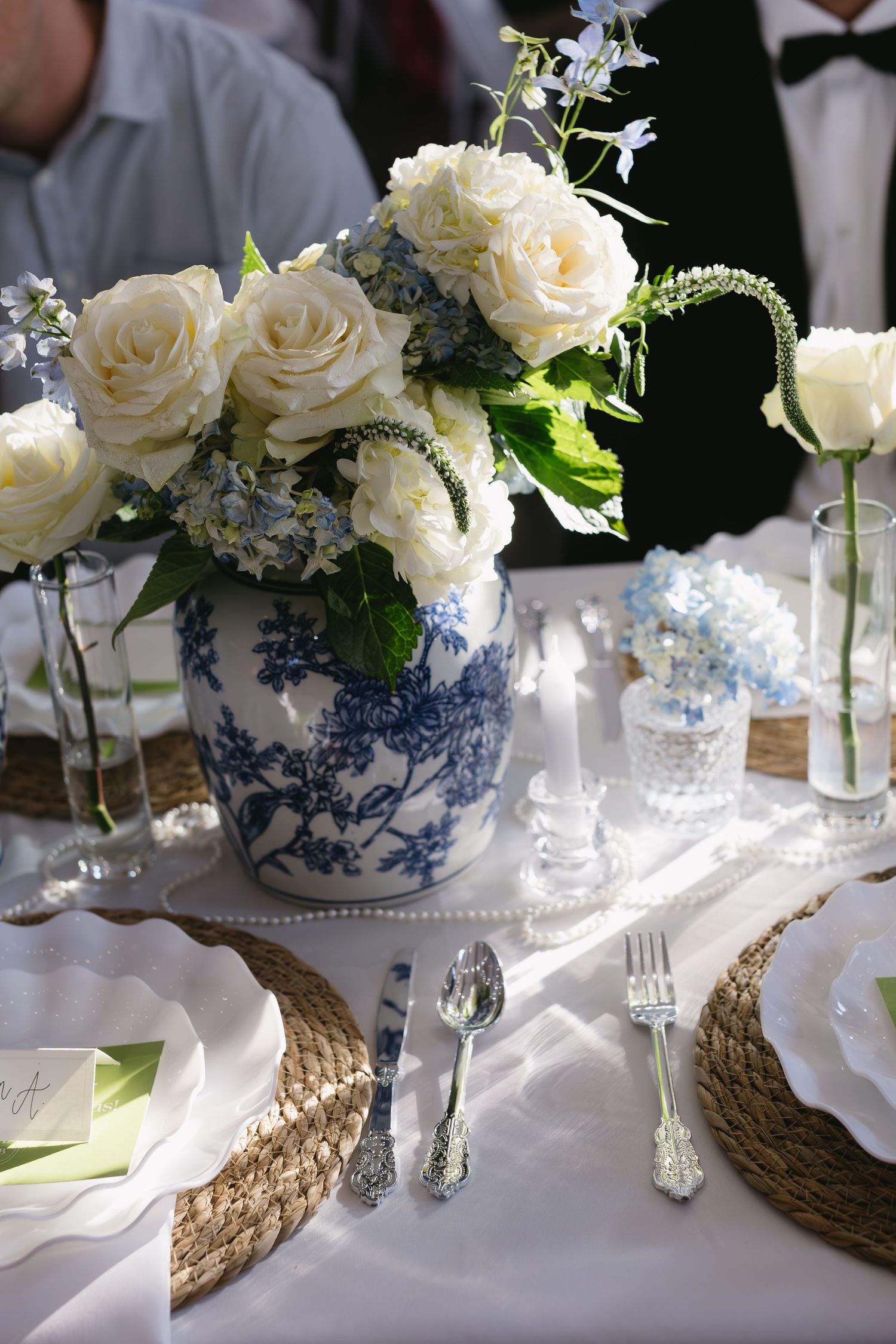 Elegant table settings with blue and white china vases holding white roses on woven charger plates.