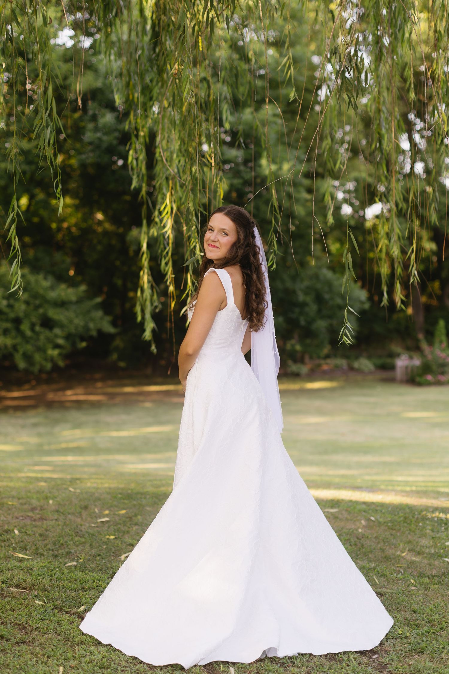 A bride in a flowing white gown stands under a weeping willow tree in a garden setting.
