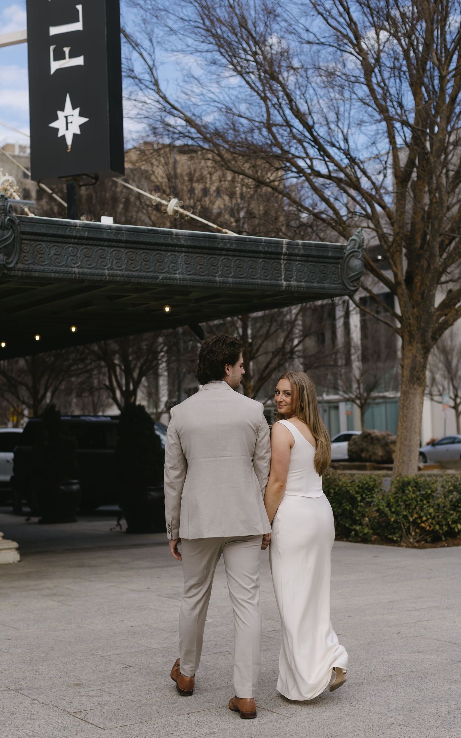 A couple in formal attire walks together on a paved outdoor area near trees.
