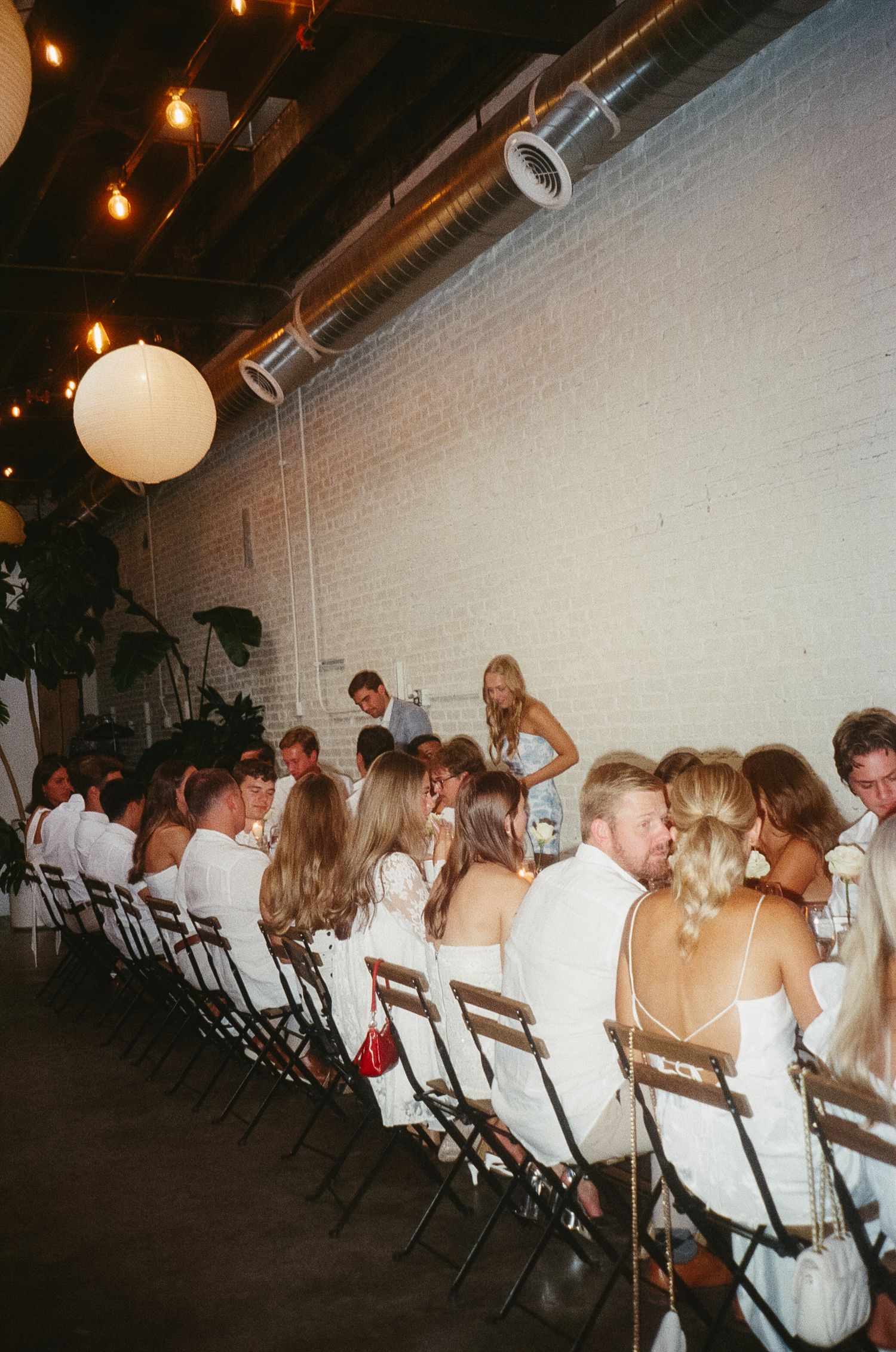 A row of wedding guests seated in white chairs at an indoor venue with string lights and white brick walls.
