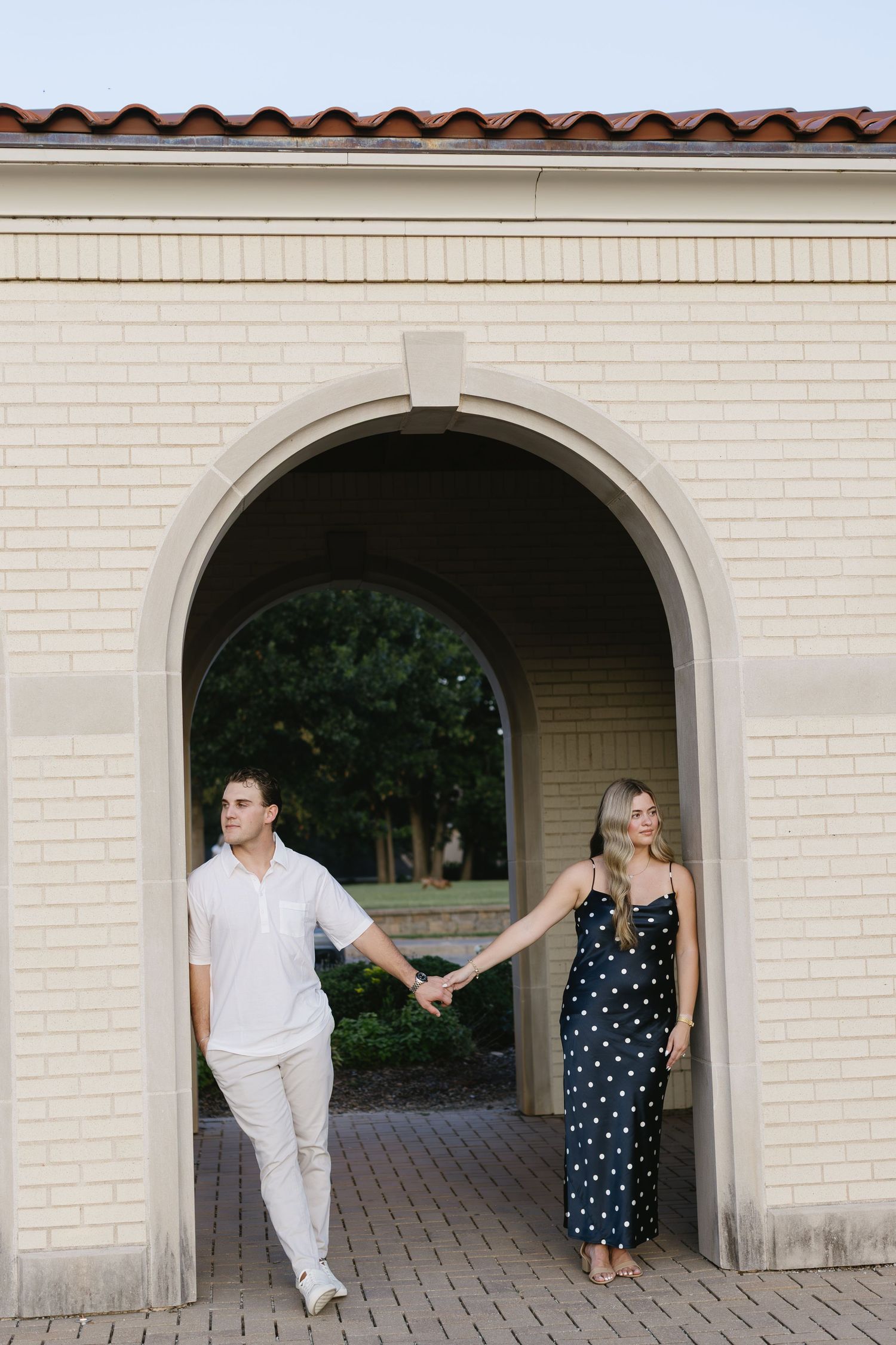 A couple holding hands while walking through a white arched doorway, her in a polka dot dress and him in light clothes.