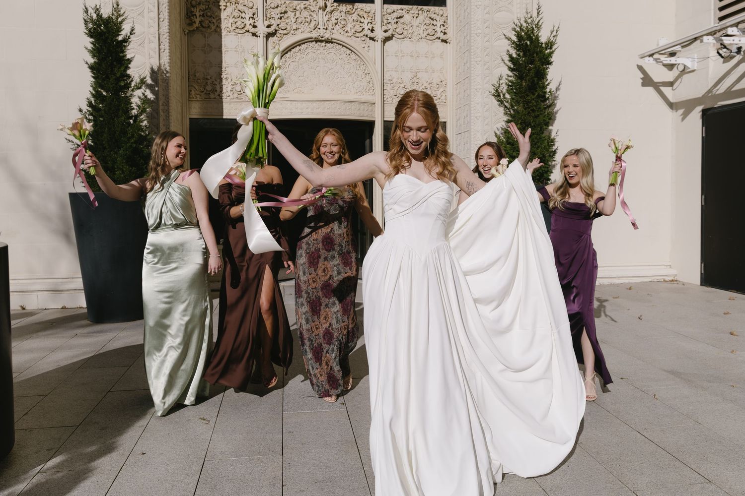 A celebratory wedding party moment outside with bridesmaids in various colored dresses holding flowers.