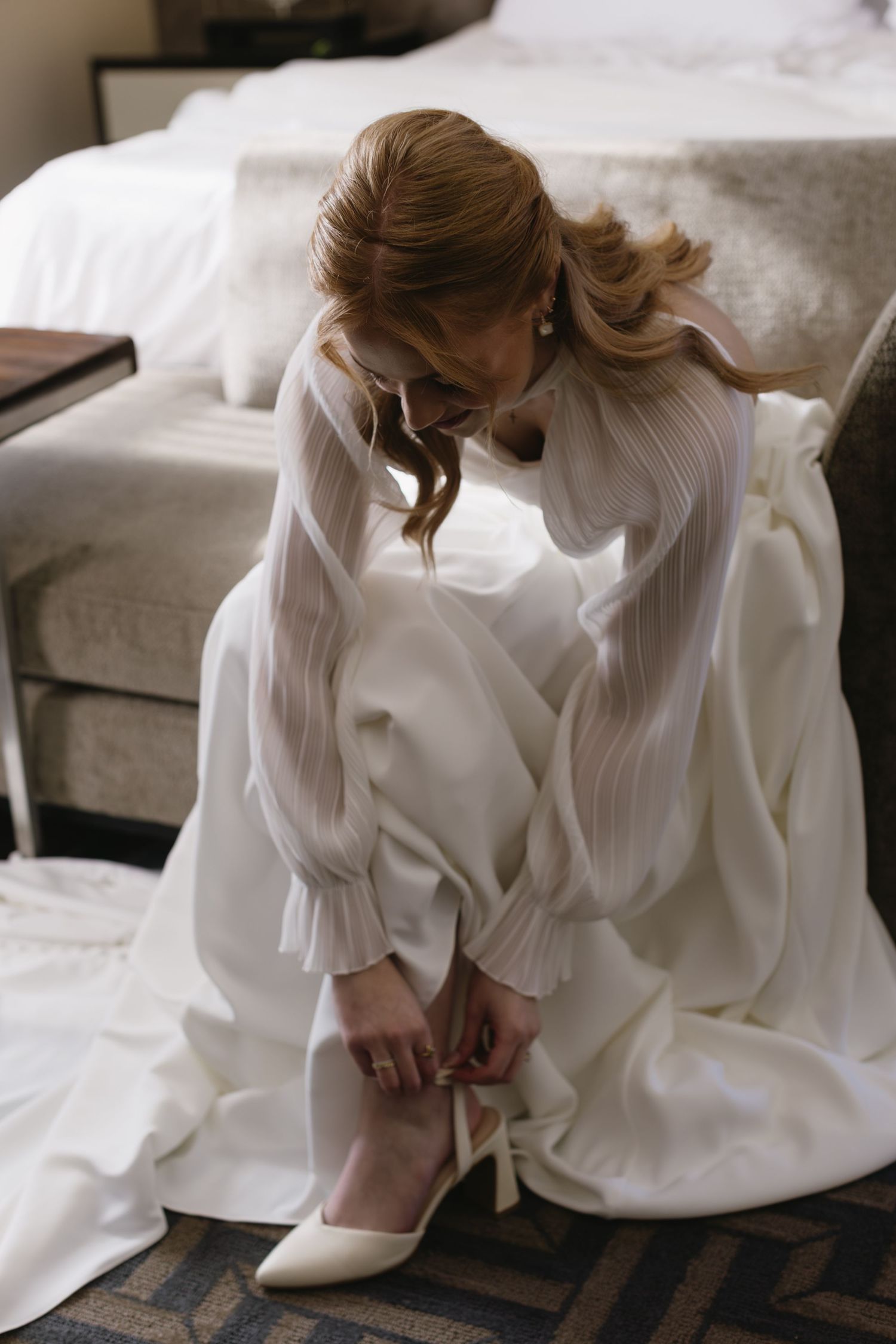 Bride in white long-sleeved dress sits gracefully on a light-colored couch.