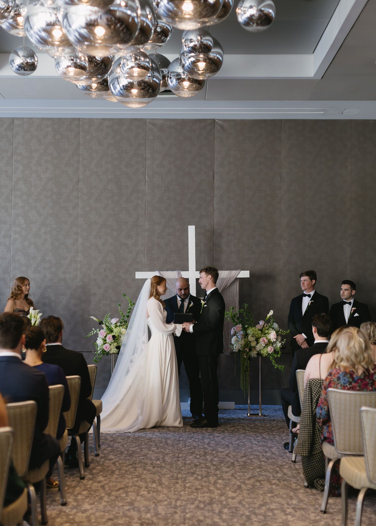 Wedding ceremony in elegant hotel ballroom with modern chandelier and cross at altar.