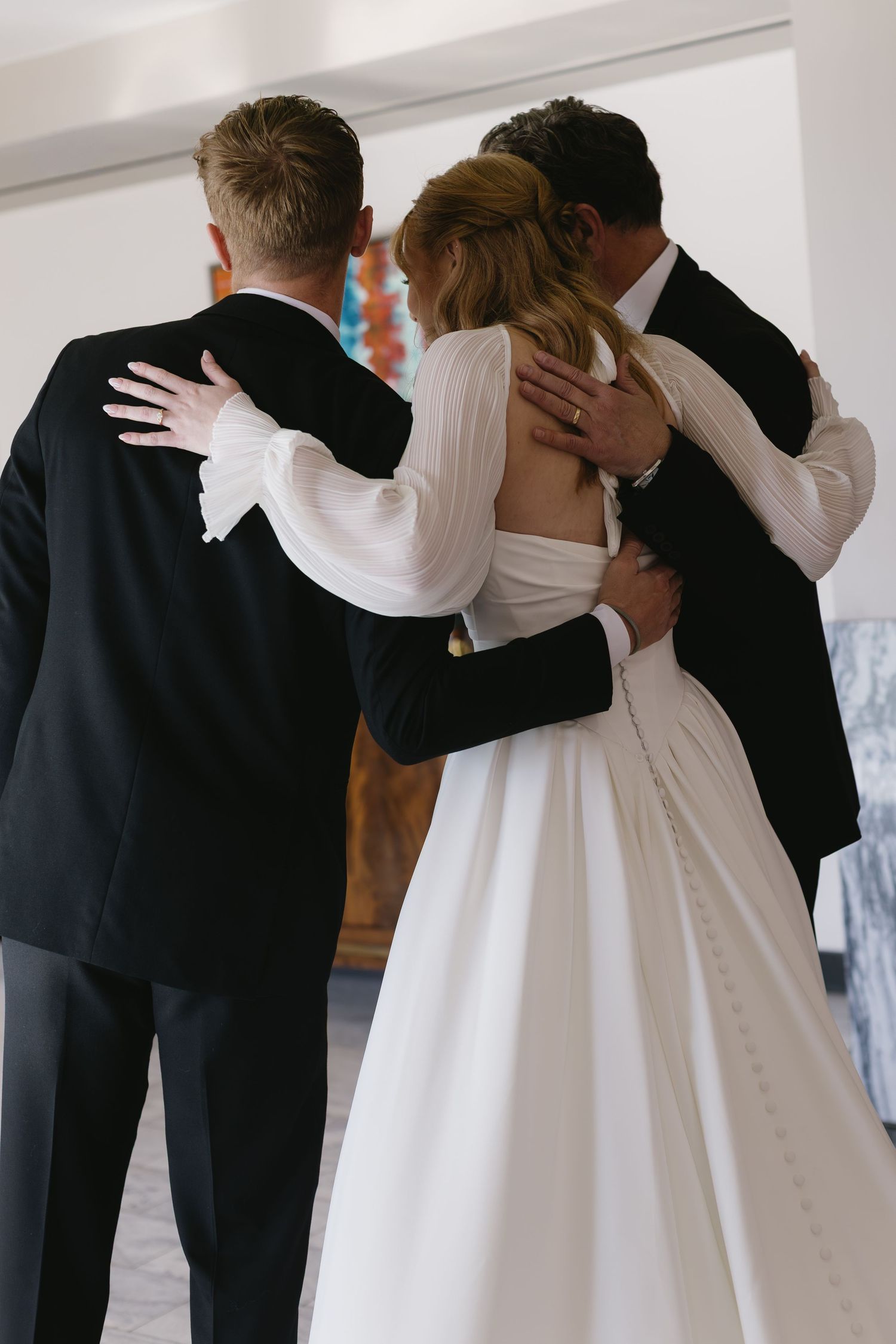 Wedding party lineup in black suits and white dress embracing in bright indoor space.
