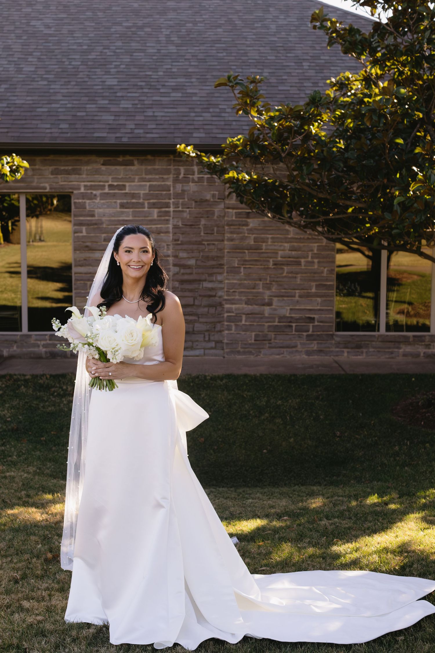 A bride in a flowing white wedding gown with a long train poses outdoors against a brick building.