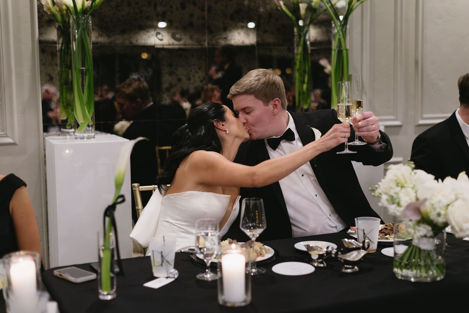 Elegant wedding reception with couple toasting at black-clothed table with white floral centerpieces.
